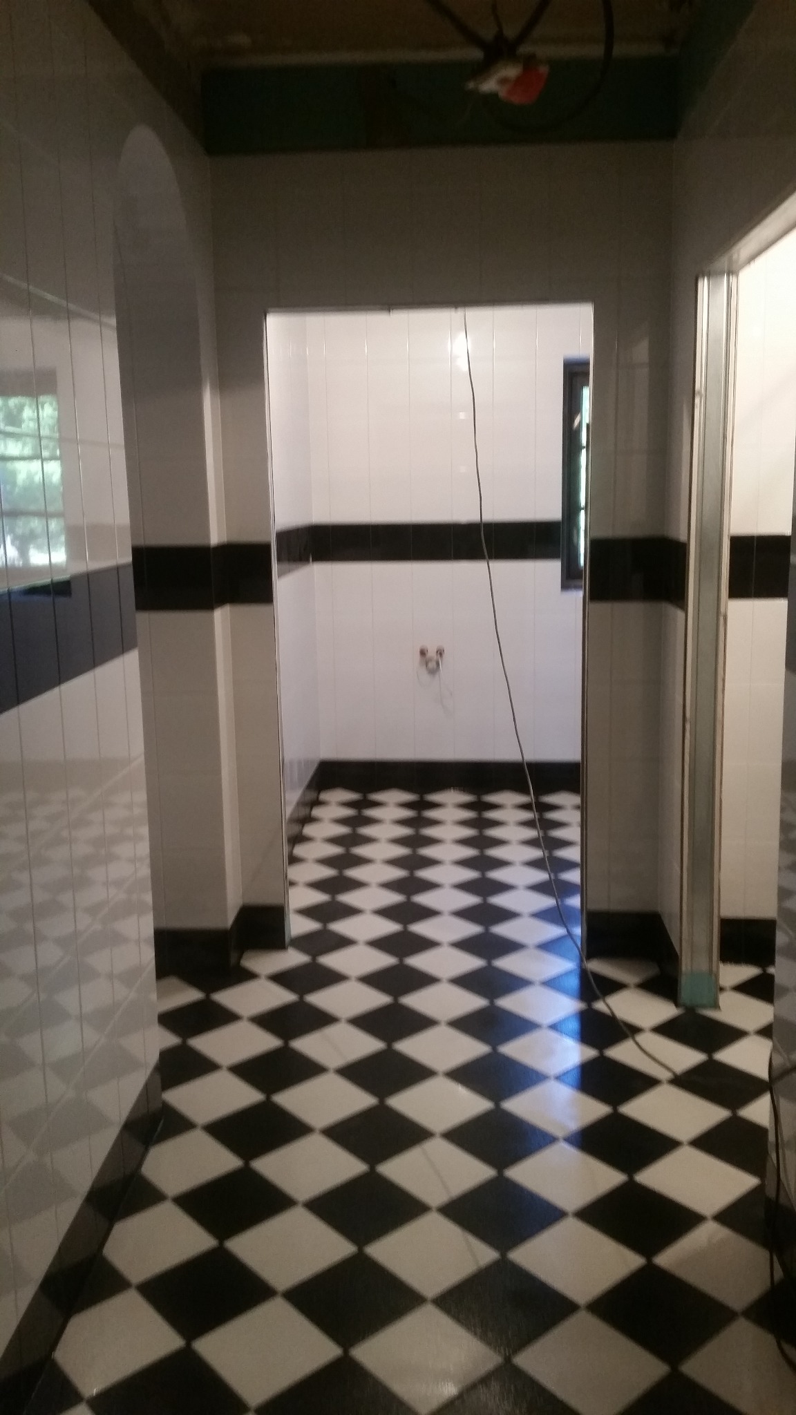 Wykończona łazienka z czarno-białą szachownicą na podłodze i białymi płytkami na ścianach z czarnym pasem. Widoczne elementy instalacji. Brak sufitu.