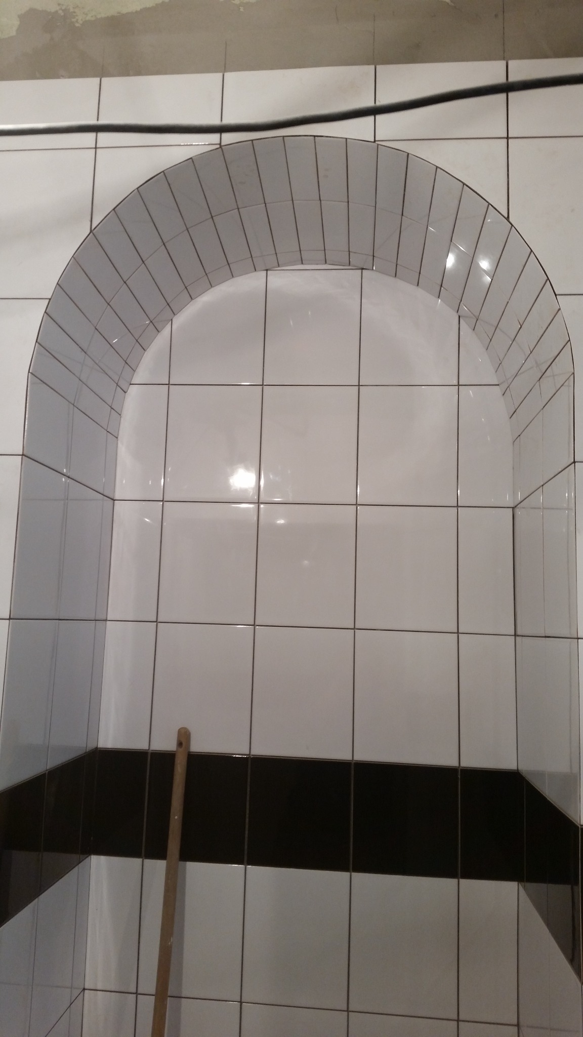 Nisza w łazience wyłożona białymi płytkami z czarnym pasem akcentującym, uformowana w łuk. Widoczny fragment drewnianego narzędzia.