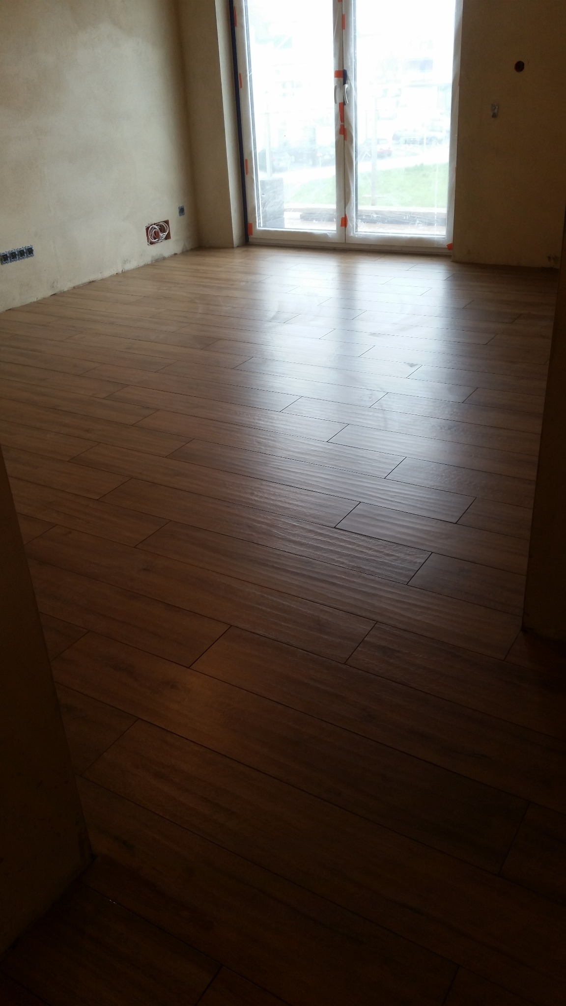 Ukończone układanie paneli podłogowych w nowym mieszkaniu, widoczne gniazdka elektryczne i okno balkonowe. Brązowe panele ułożone równo, ściany przygotowane do malowania.