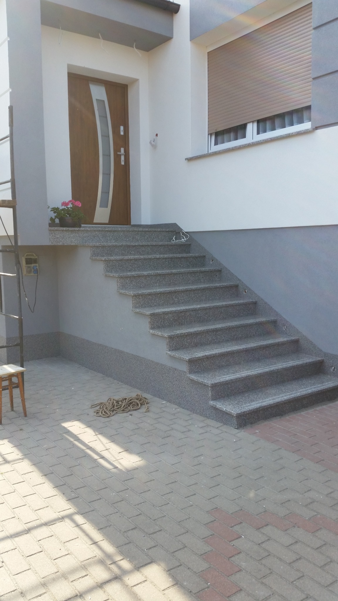 Granitowe schody wejściowe do domu z drewnianymi drzwiami i roletami w oknie. Widoczne narzędzia i kostka brukowa.