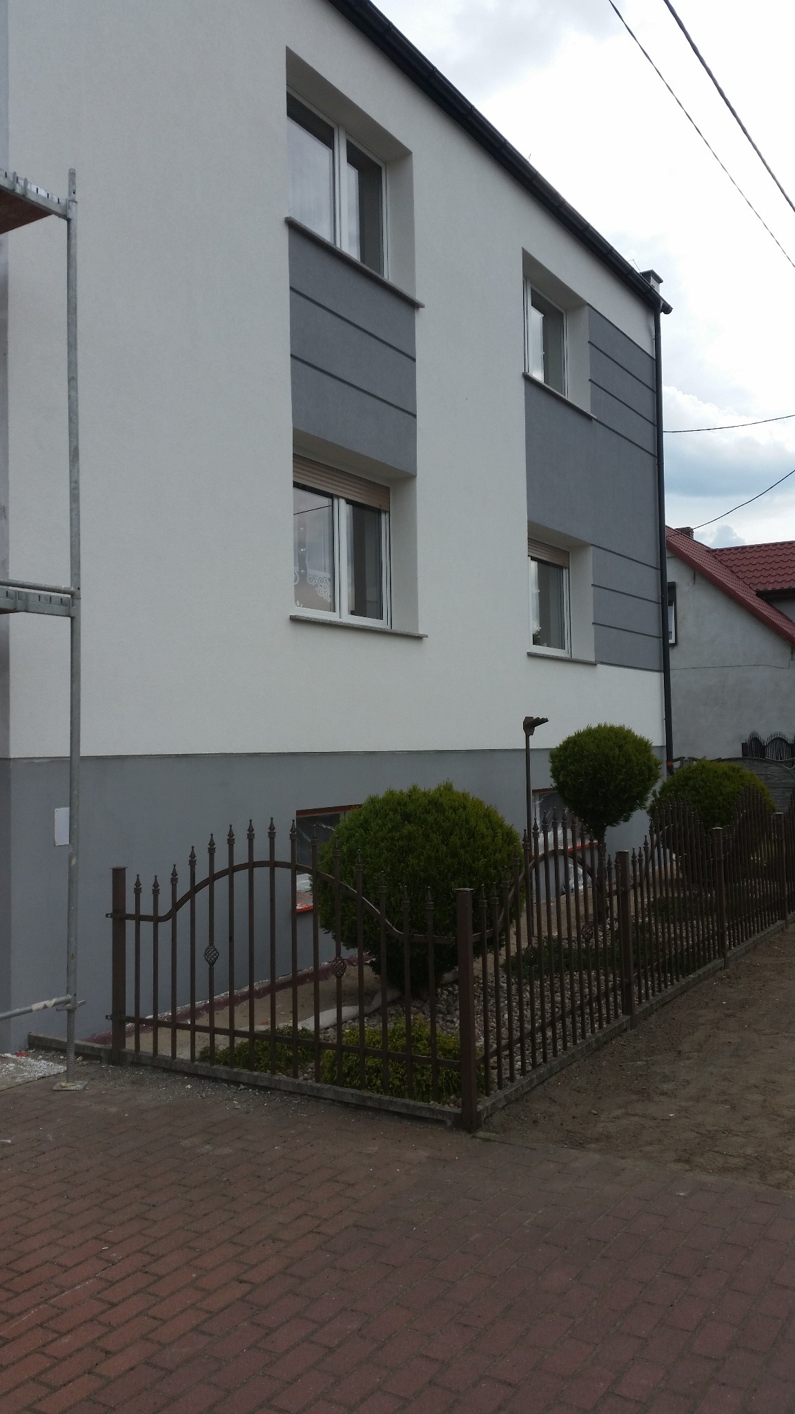 Ocieplony budynek z szarą elewacją i brązowym ogrodzeniem. Fragment rusztowania po lewej. Widoczne krzewy i kostka brukowa. Ujęcie z dołu, podkreślające wysokość budynku.