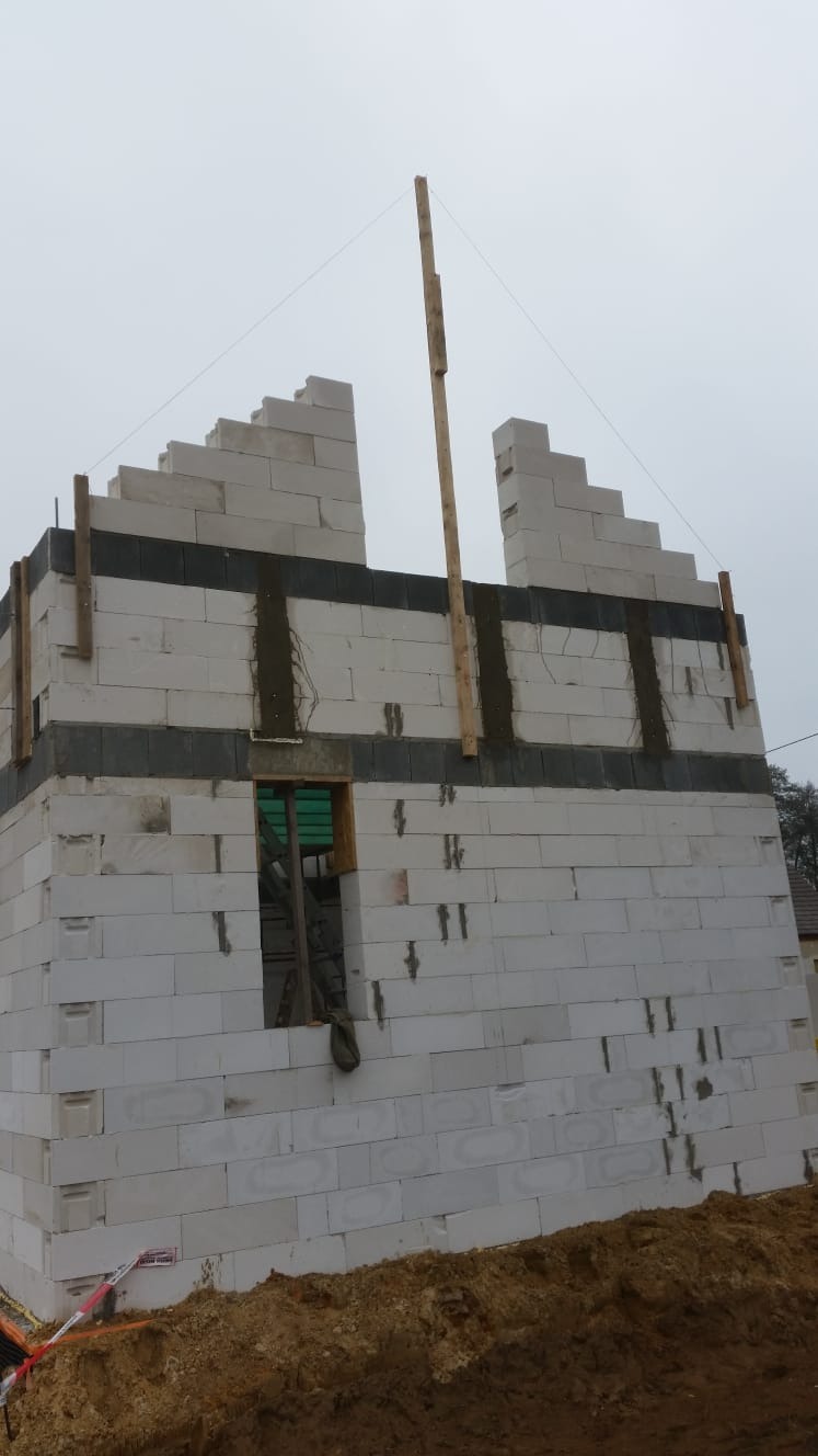 Budowa domu z białych bloczków, widoczne drewniane stemple i sznurki wyznaczające pion, okno z zieloną folią zabezpieczającą, etap wznoszenia ścian.