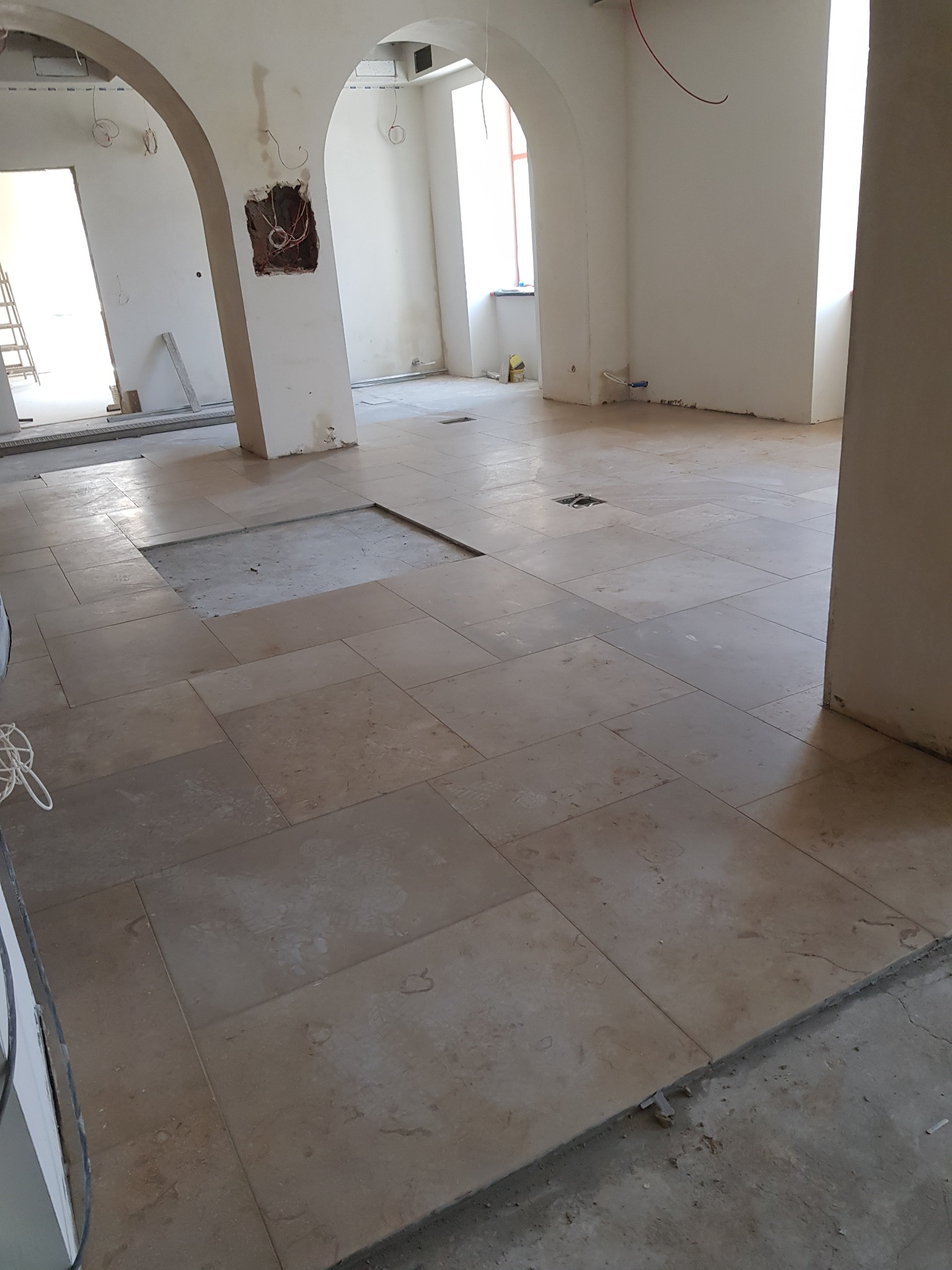 Jasne płytki podłogowe w przestronnym wnętrzu z łukowymi przejściami, częściowo ułożone, z widoczną instalacją elektryczną w ścianie. Prace wykończeniowe w toku.
