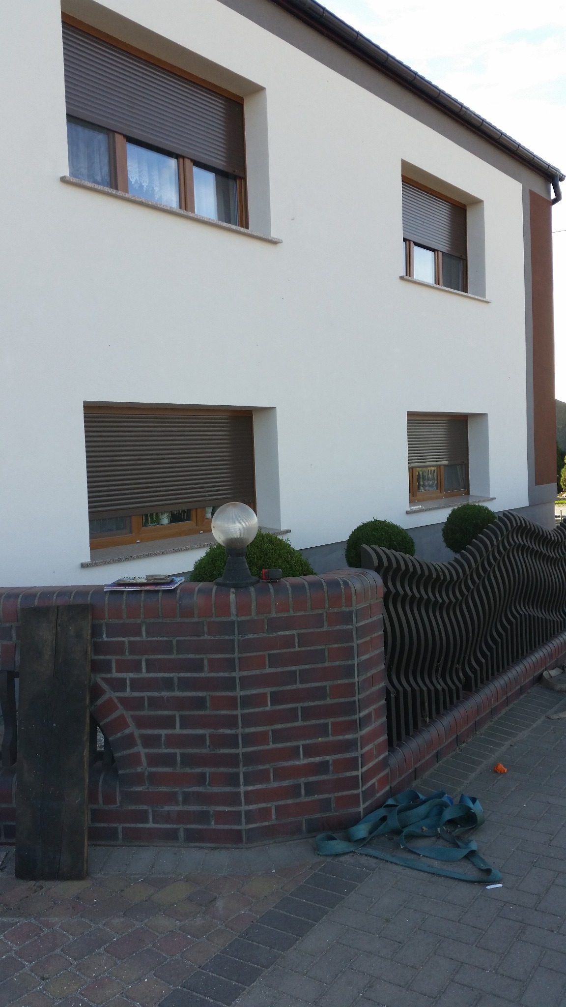 Biała fasada domu z brązowymi roletami w oknach. Czerwona ceglana podmurówka z ozdobnym metalowym ogrodzeniem i okrągłą lampą. Brukowany chodnik i pasy transportowe.