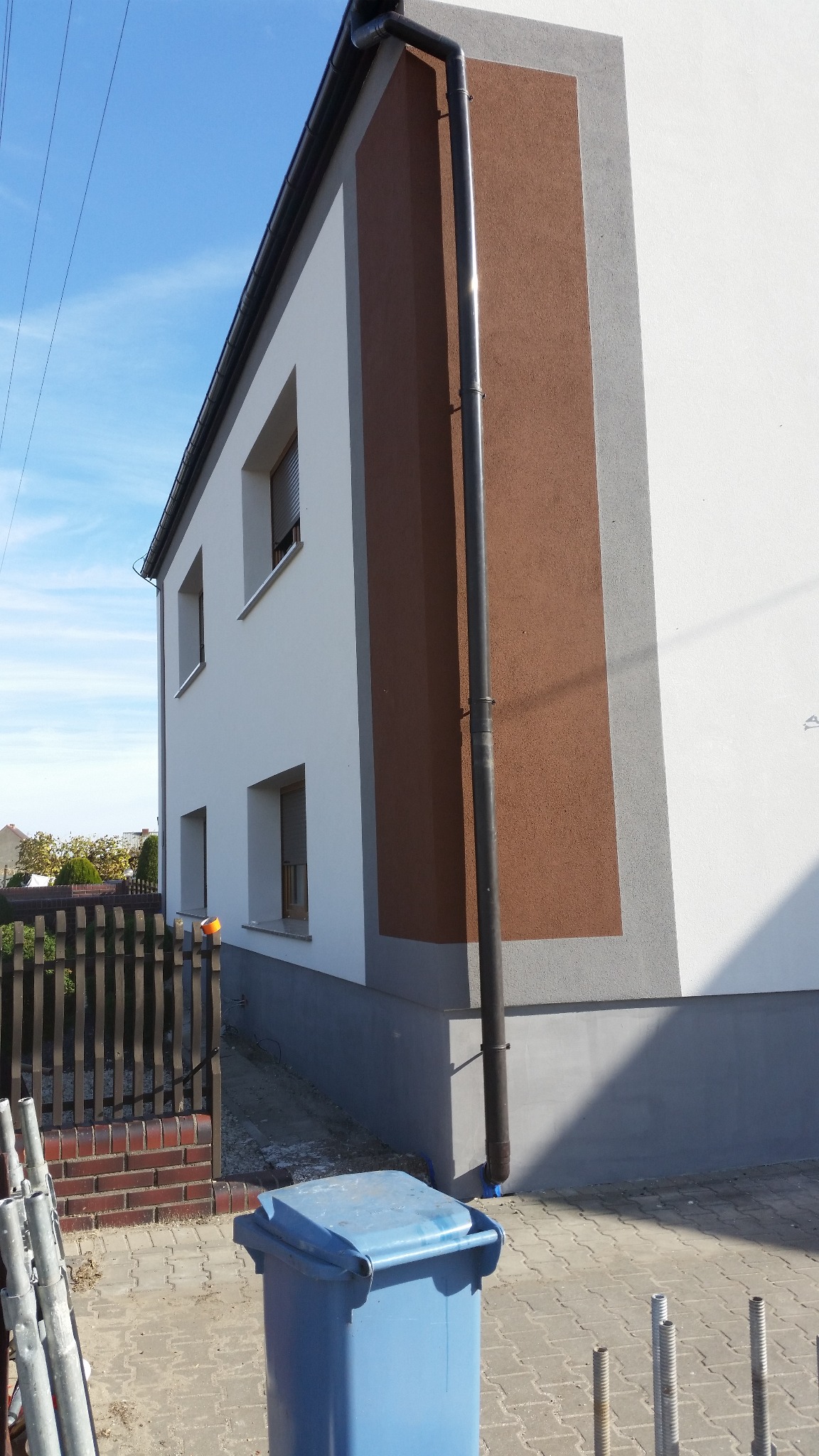Dwukondygnacyjny dom po ociepleniu elewacji, z brązowym pasem i szarymi akcentami. Widoczna rynna, fragment ogrodzenia i niebieski kosz na śmieci.