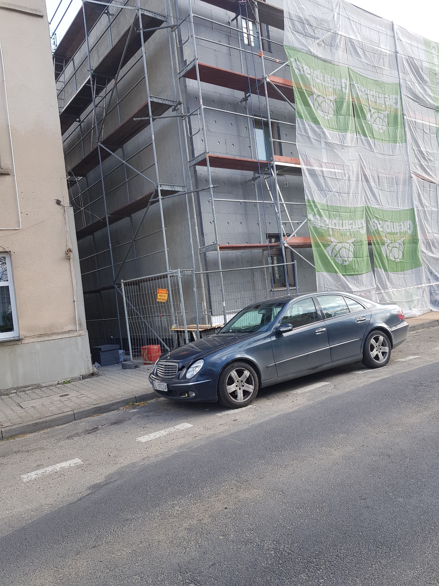 Budynek w rusztowaniach, zabezpieczony siatką z reklamą, obok zaparkowany samochód. Trwa etap ocieplania ścian zewnętrznych.