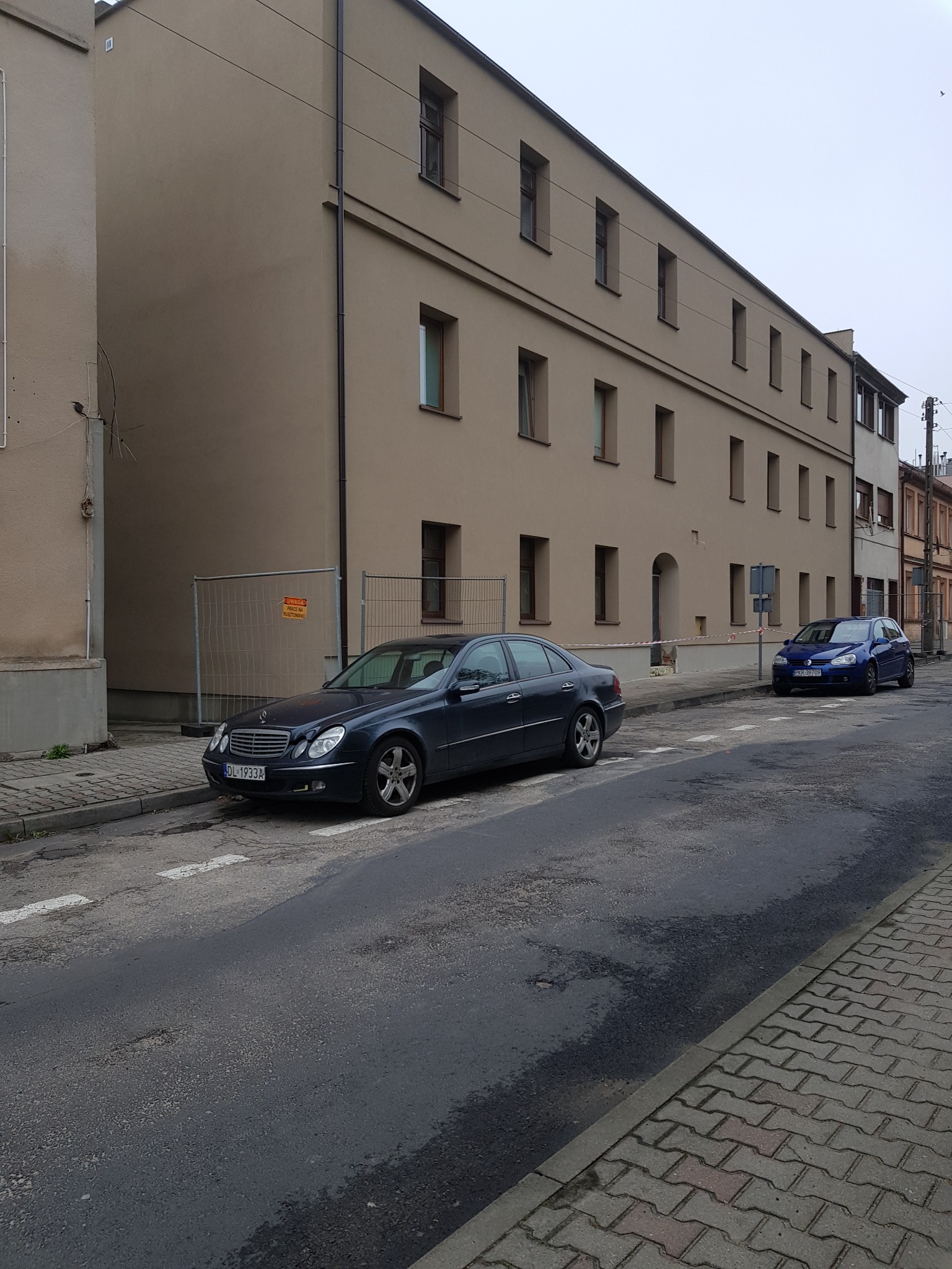 Budynek po renowacji elewacji w odcieniach beżu, z widocznymi dwoma samochodami na ulicy. Uwagę zwraca siatka zabezpieczająca teren prac.