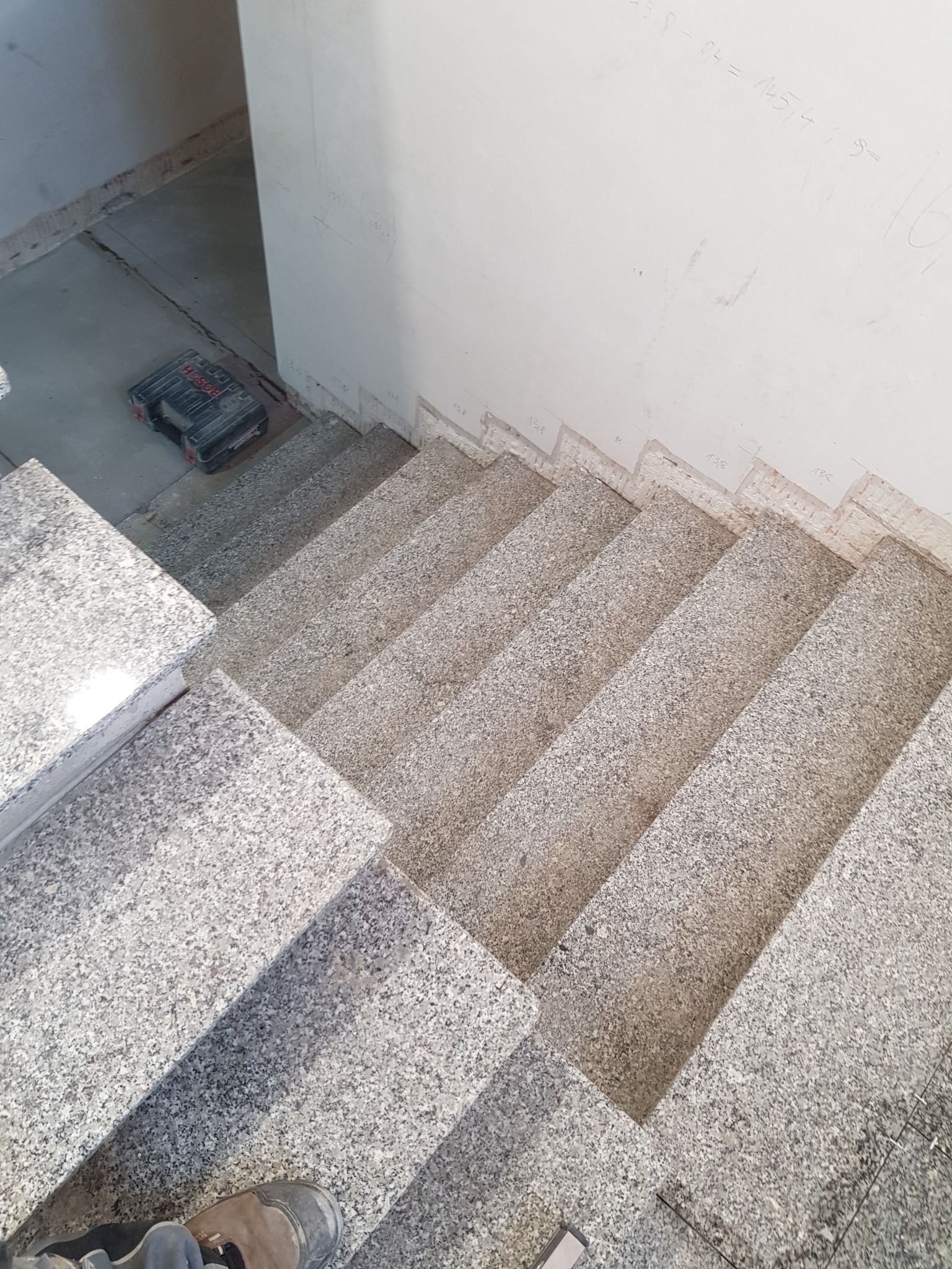 Wykończone granitowe schody wewnętrzne z widocznymi śladami po pomiarach na ścianie, skrzynka narzędziowa w tle. Widok z góry, fragmentarycznie widoczne buty.