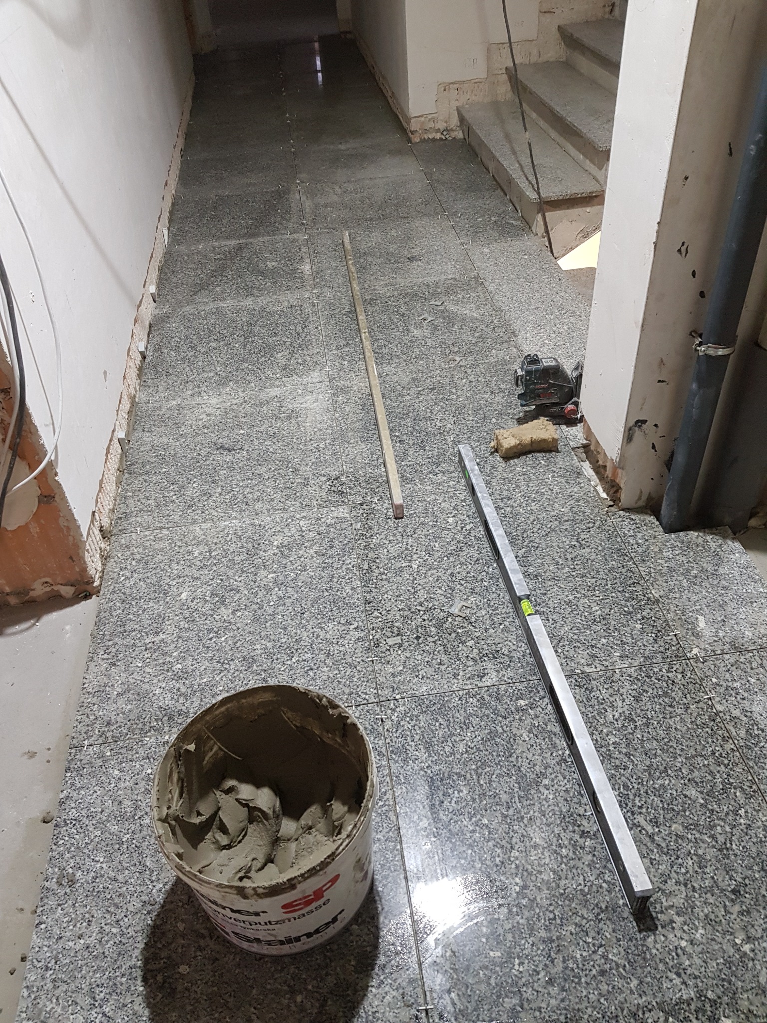 Układanie szarej, nakrapianej terakoty w korytarzu. Widoczne narzędzia: poziomica, laser krzyżowy, wiadro z zaprawą. Etap prac: częściowo ułożone płytki, pozostałe do ułożenia.