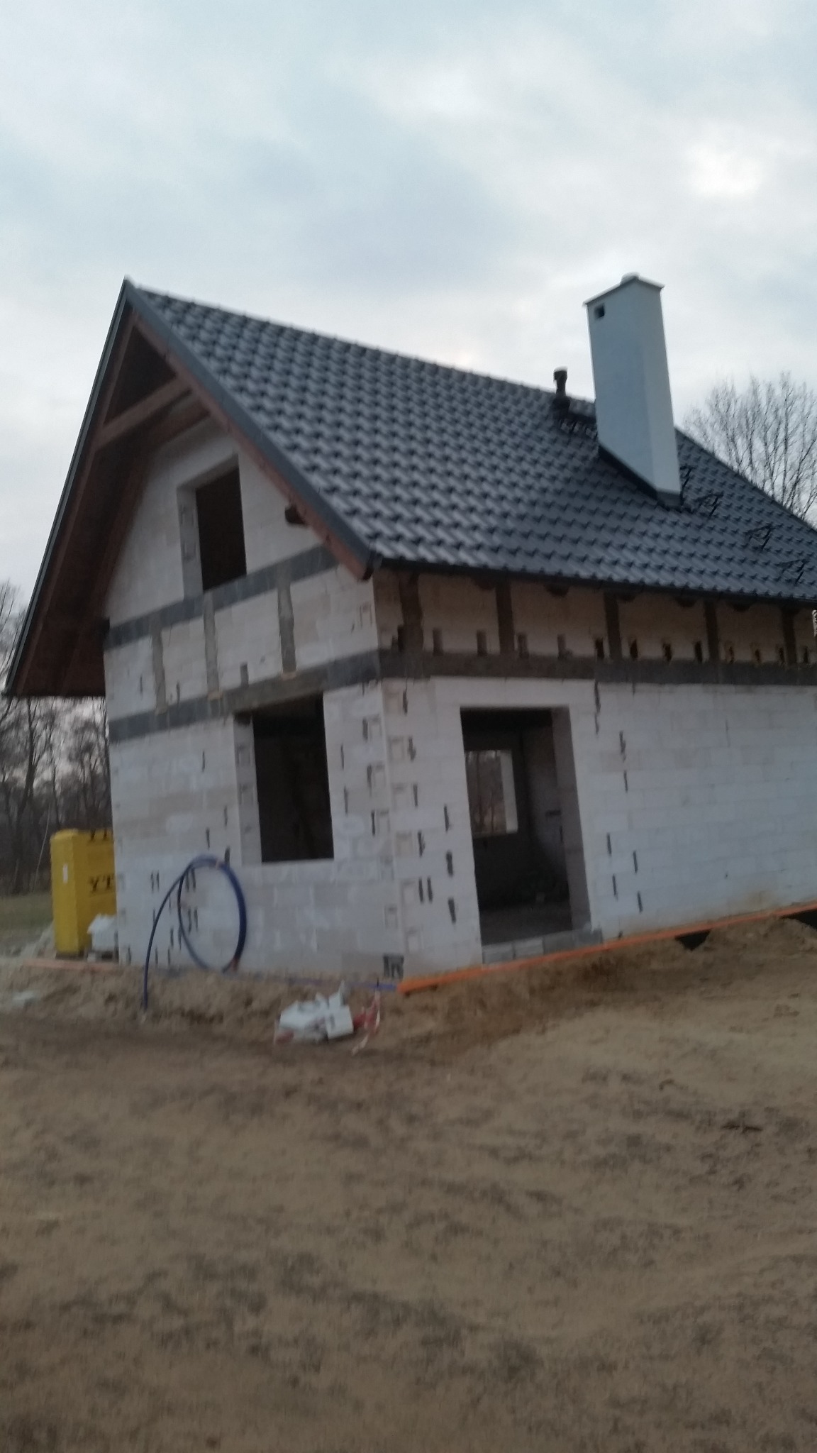 Surowy stan domu jednorodzinnego z ciemną dachówką i kominem, widoczne otwory okienne i drzwiowe, żółta kostka izolacyjna obok budynku.