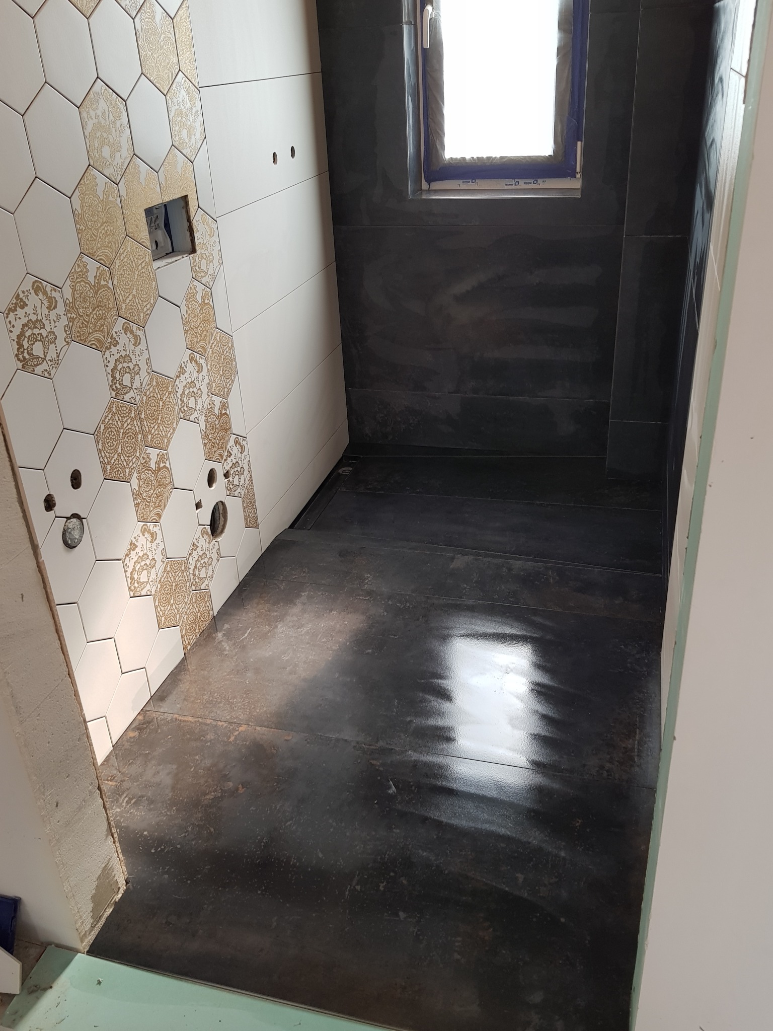 Łazienka w trakcie wykończenia: heksagonalne płytki z motywem kwiatowym na ścianie, ciemna podłoga i okno. Widoczne przygotowane otwory na armaturę.