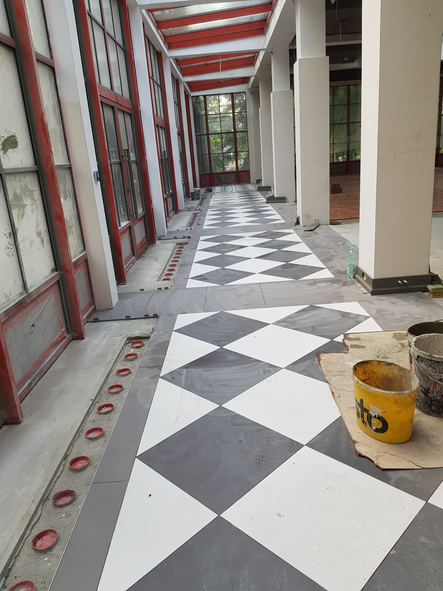 Układanie podłogi z płytek w szachownicę w korytarzu z oknami. Widoczne narzędzia i fragment podłogi przed położeniem płytek. Prace wykończeniowe w toku.