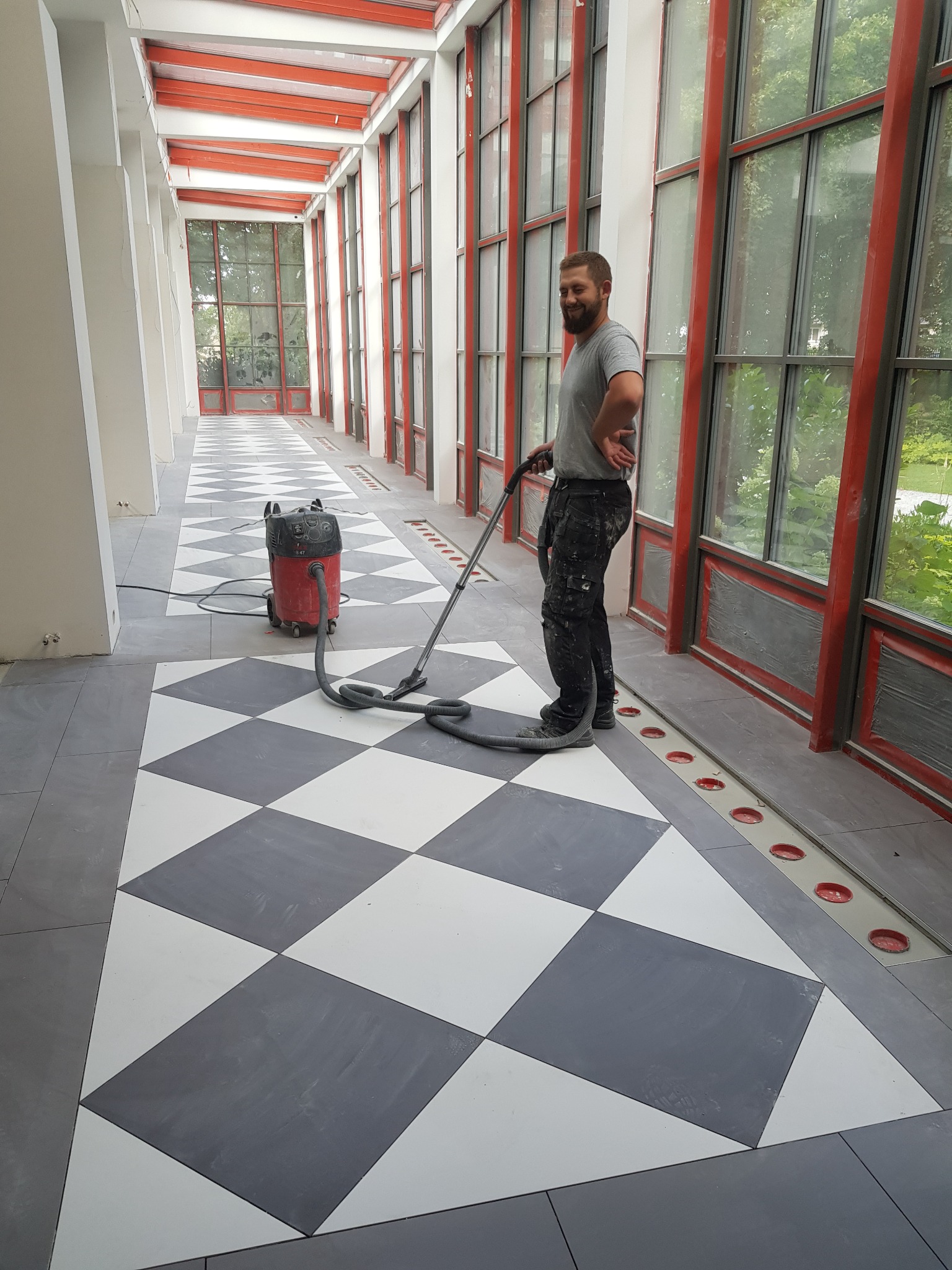 Pracownik z odkurzaczem przemysłowym czyści nową, biało-szarą, szachownicową posadzkę w przeszklonym korytarzu z czerwonymi ramami okiennymi.