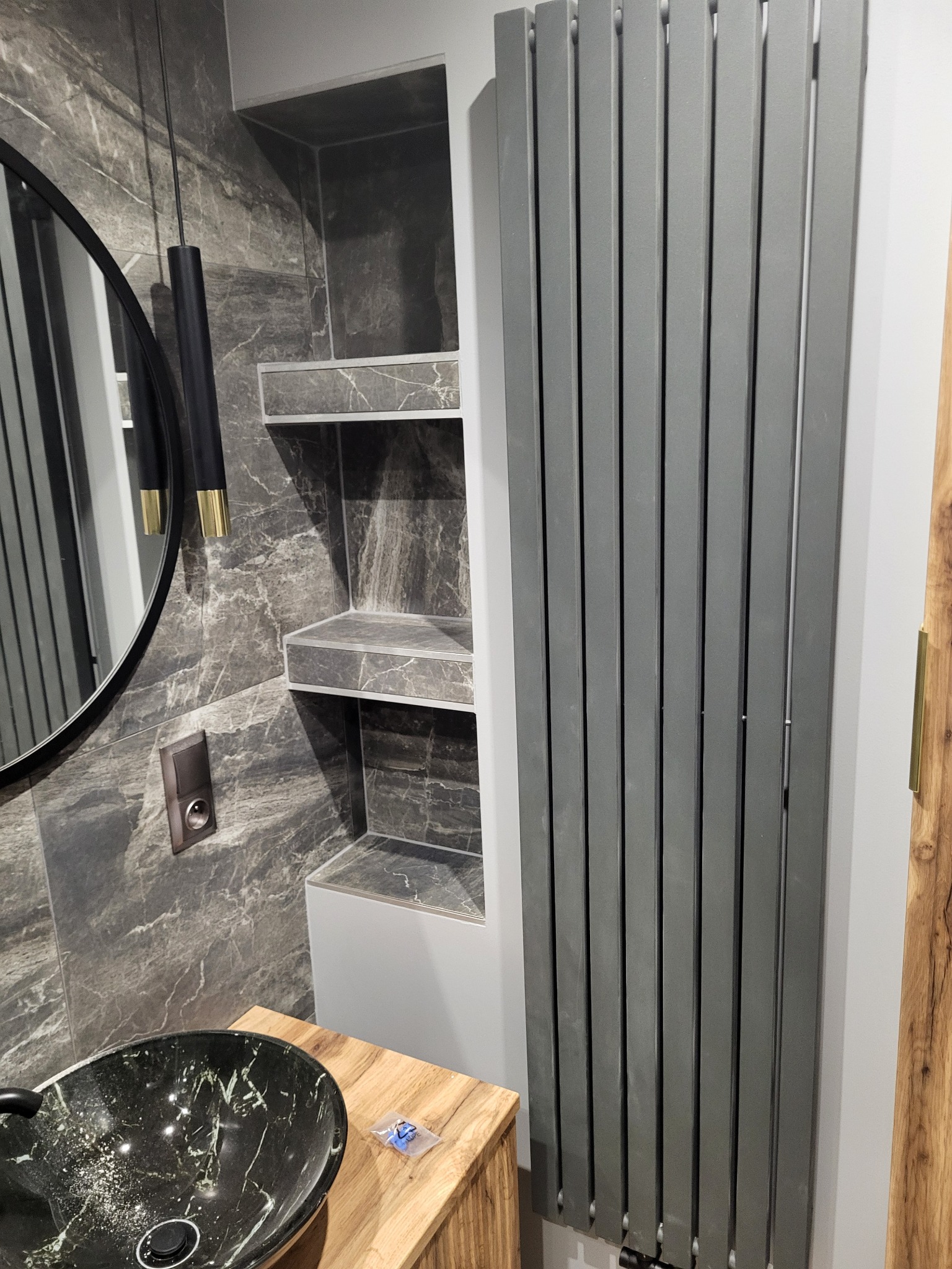 Nowoczesna łazienka z umywalką w kolorze czarnym z zielonymi żyłkami, drewnianą szafką, szarym grzejnikiem panelowym i marmurowymi płytkami na ścianie z wnęką z półkami.