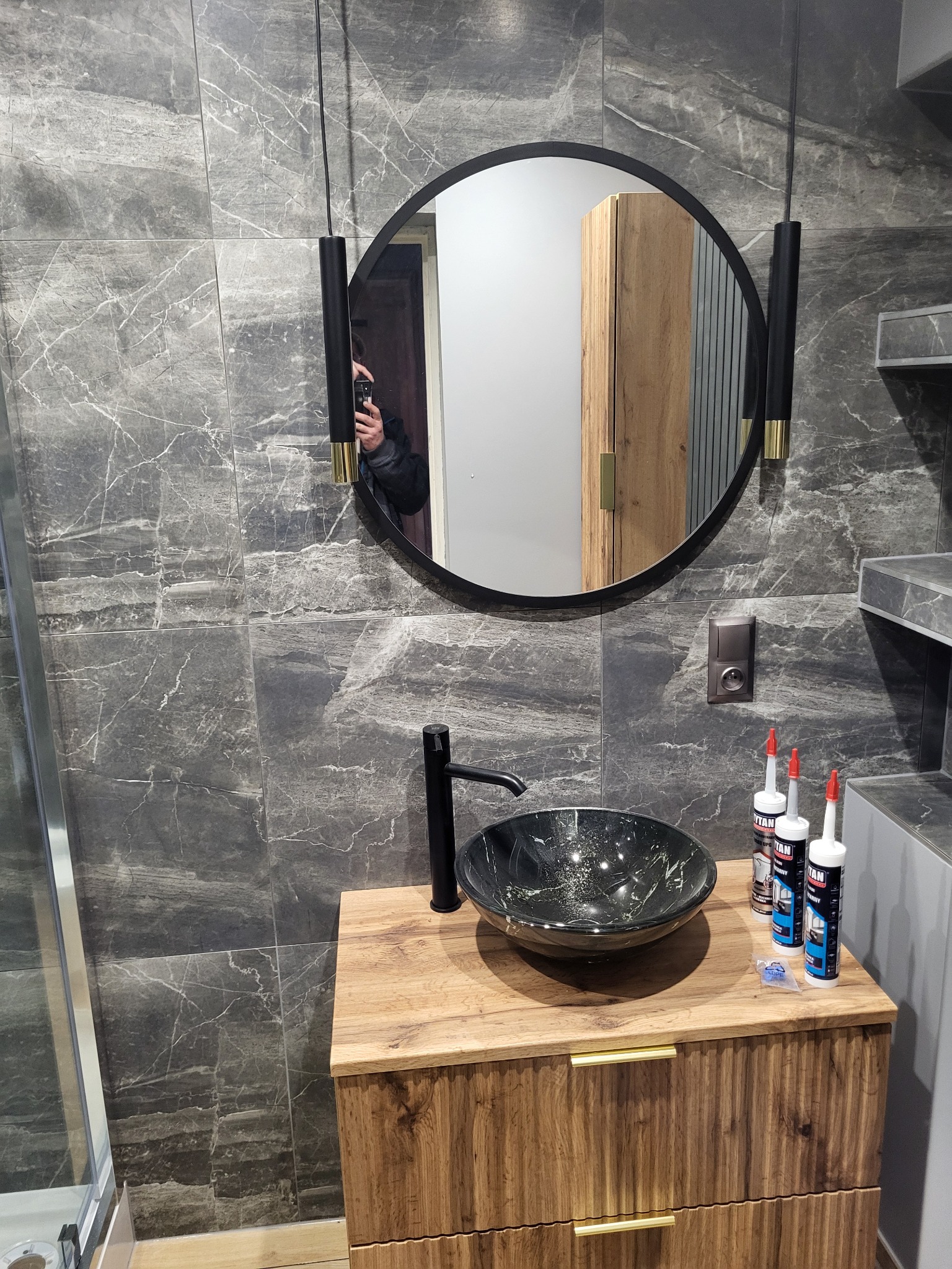 Nowoczesna łazienka z umywalką nablatową w kolorze czarnym, okrągłym lustrem z podświetleniem i szarymi płytkami imitującymi marmur na ścianach, widoczne tuby silikonu na blacie.