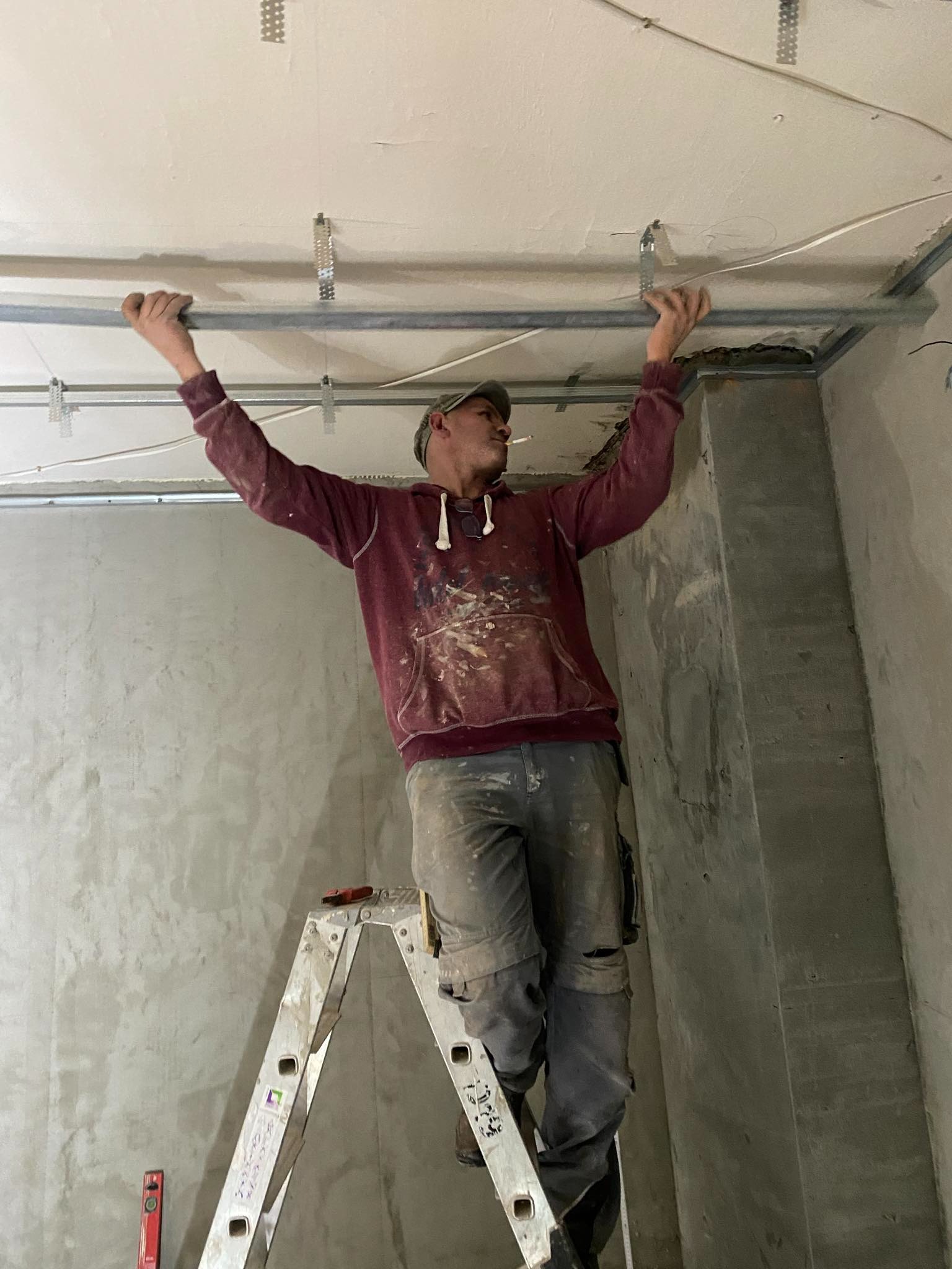 Mężczyzna na drabinie, w ubrudzonym farbą swetrze i czapce, montuje metalowy stelaż pod sufitem, trzymając w ustach papierosa. Ściany surowe, w tle widoczny pionowy słup. Na szczeblach drabiny...