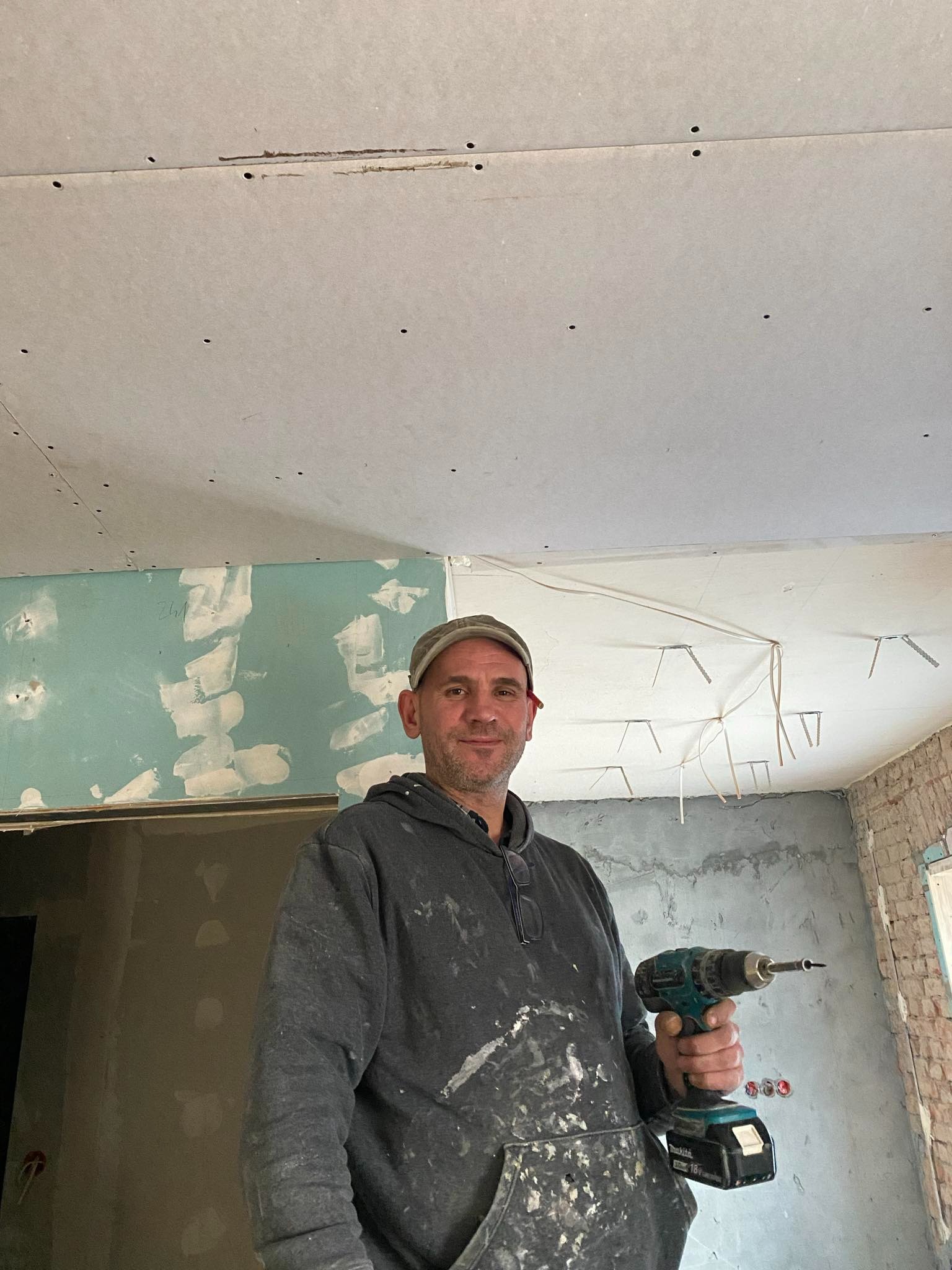 Mężczyzna w ubrudzonej bluzie trzyma wiertarko-wkrętarkę na tle sufitu z zamontowanymi płytami gipsowo-kartonowymi i fragmentów ścian w trakcie prac wykończeniowych.