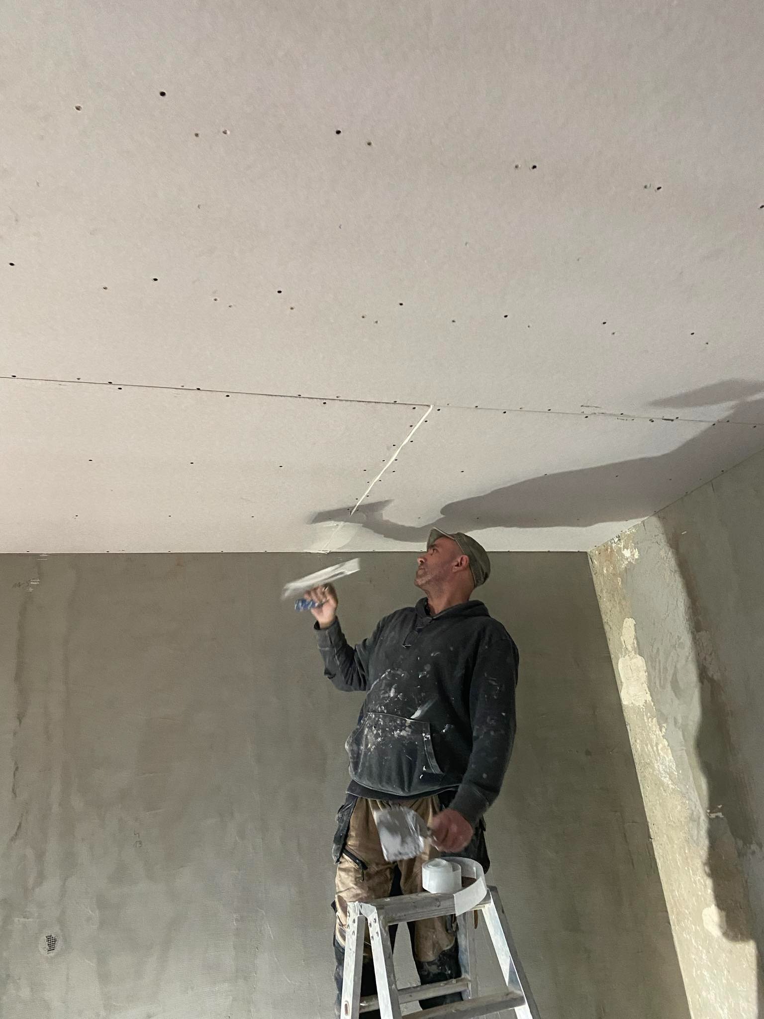 Mężczyzna na drabinie szpachluje łączenie płyt na suficie, widoczne otwory po wkrętach w płycie, ściany surowe, w tle narożnik pokoju.