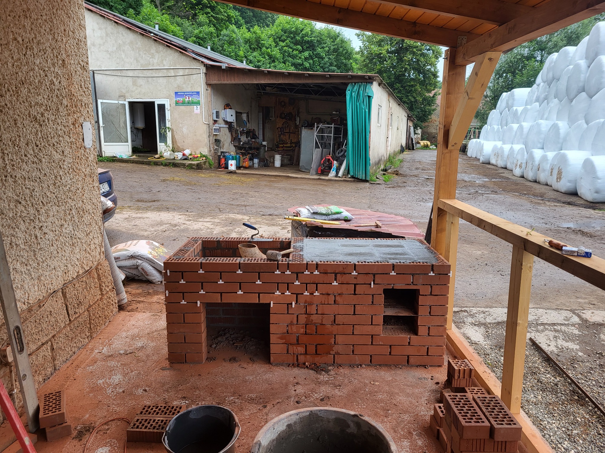 Budowa murowanego grilla z czerwonej cegły, widoczne białe krzyżyki dystansowe, zaplecze gospodarcze w tle.