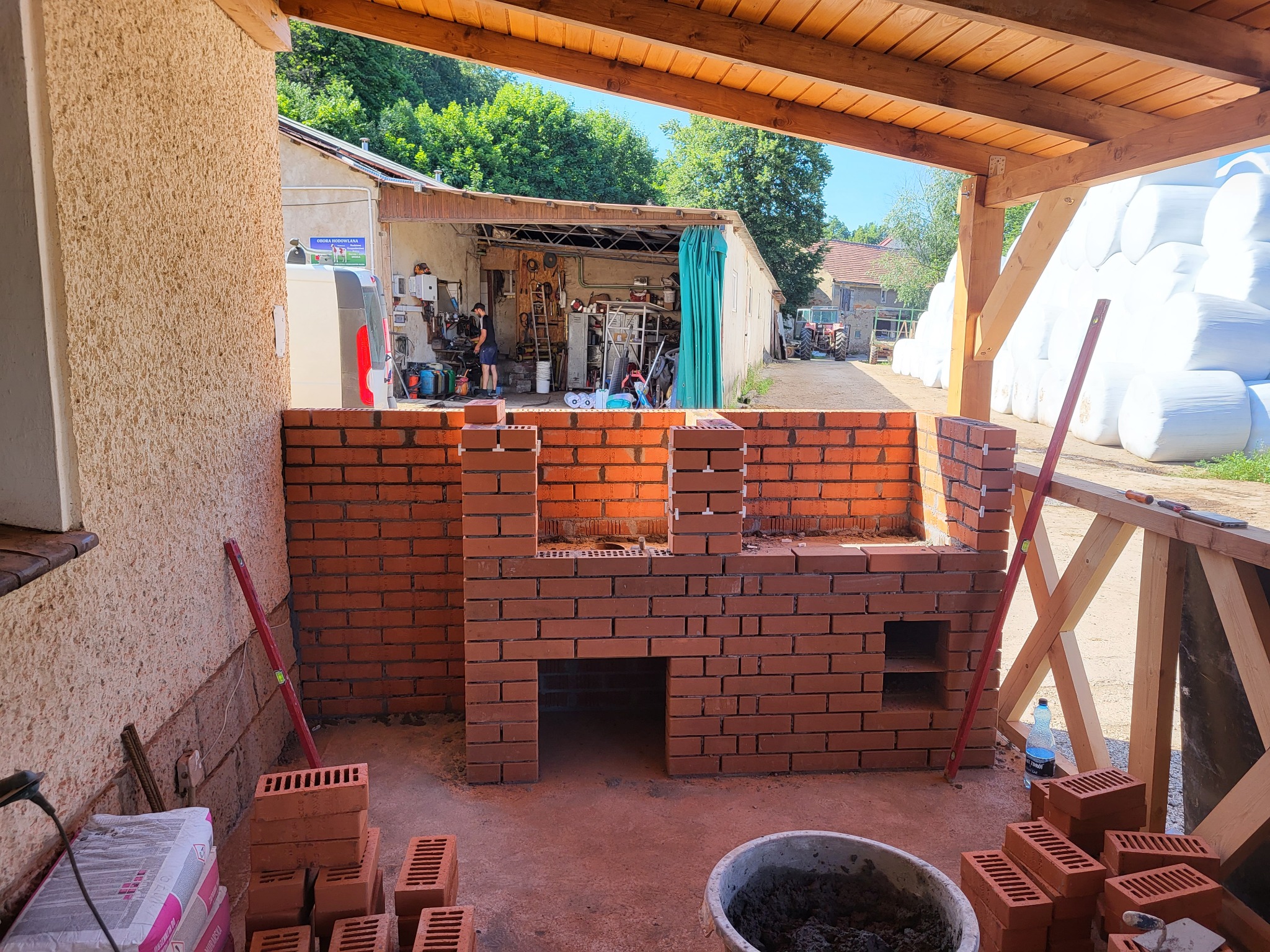Wznoszenie murowanego grilla z czerwonej cegły pod drewnianą konstrukcją z widokiem na podwórze z budynkami gospodarczymi i stertą bel siana w białej folii.