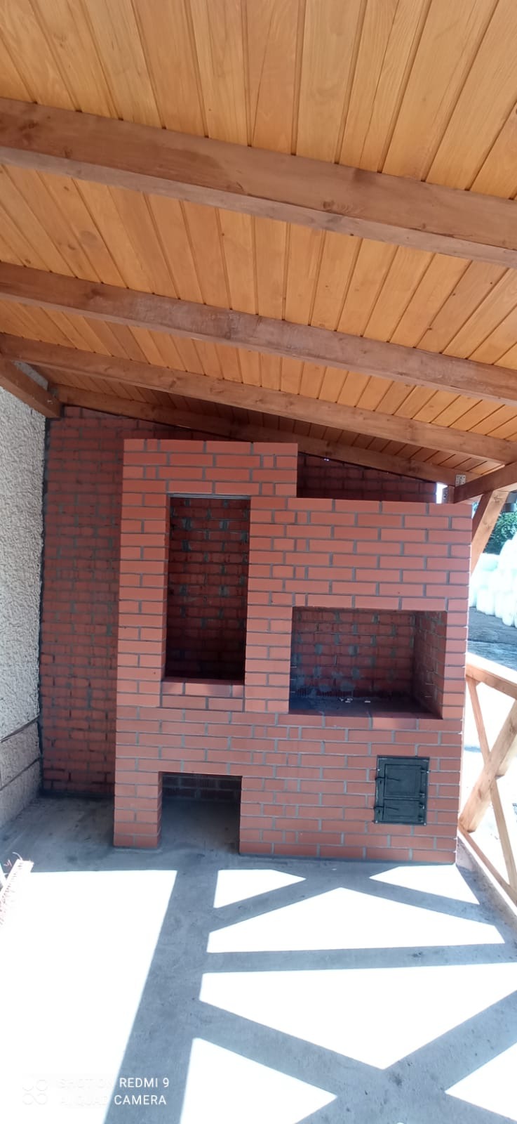 Murowany grill z czerwonej cegły pod drewnianą zadaszoną konstrukcją, z widocznym cieniem na betonowej posadzce.