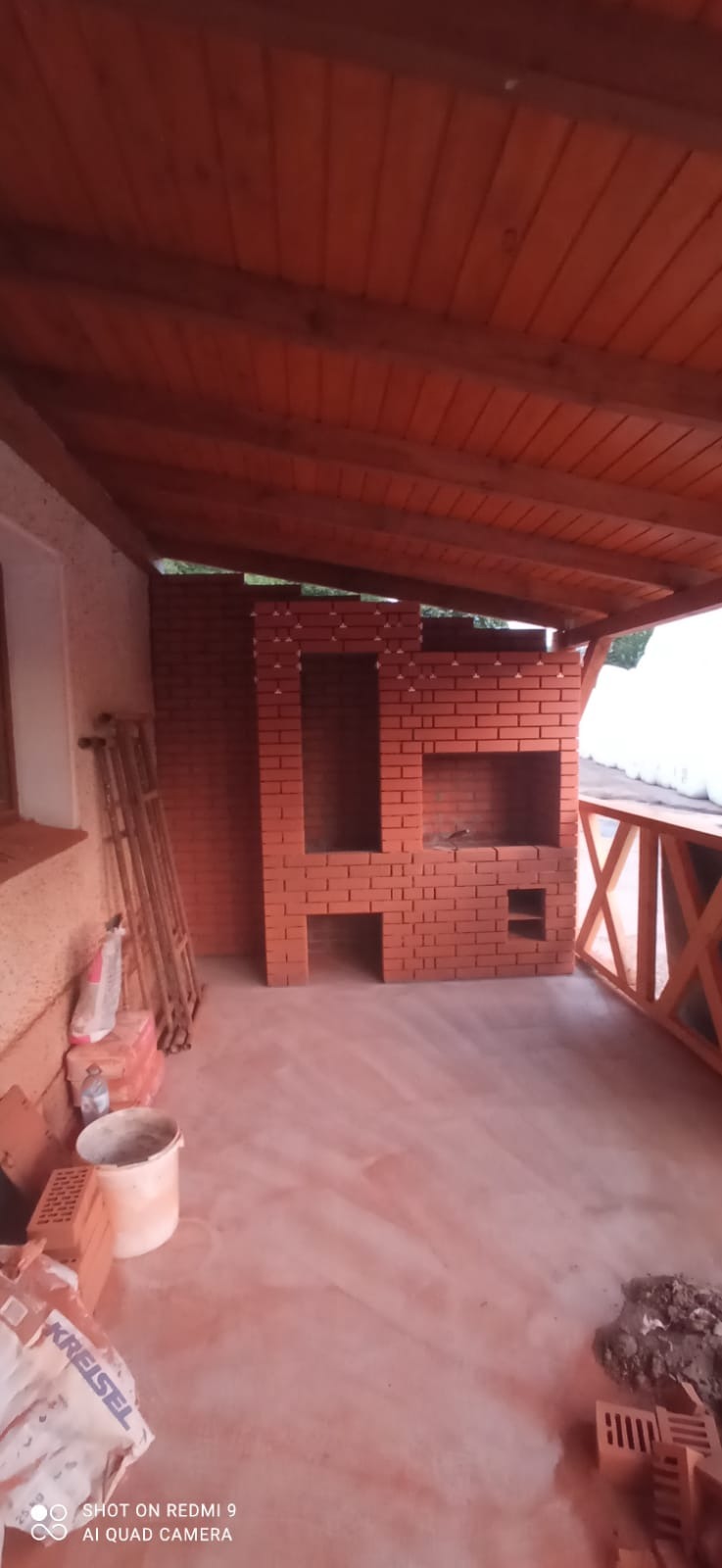 Wnętrze zadaszonego tarasu z ceglanym grillem w trakcie budowy, widoczne materiały budowlane, drewniana konstrukcja dachu i balustrada.