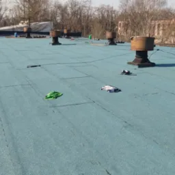 Niebieski dach pokryty papą z widocznymi kominami wentylacyjnymi, narzędziami i szmatkami w tle budynki i drzewa.