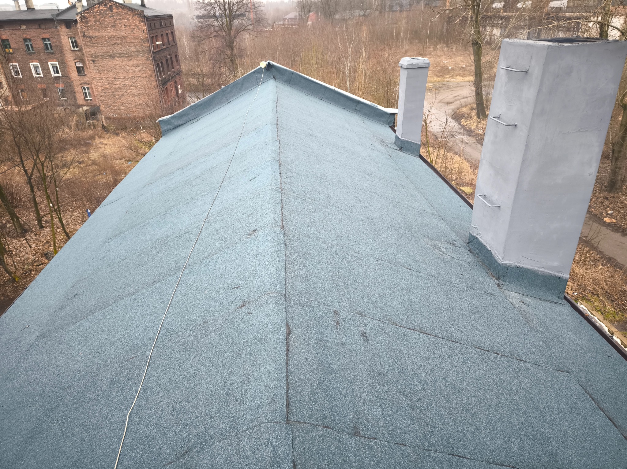 Dach pokryty papą termozgrzewalną w kolorze szaro-niebieskim, z dwoma kominami w tle i budynkiem z czerwonej cegły w oddali.