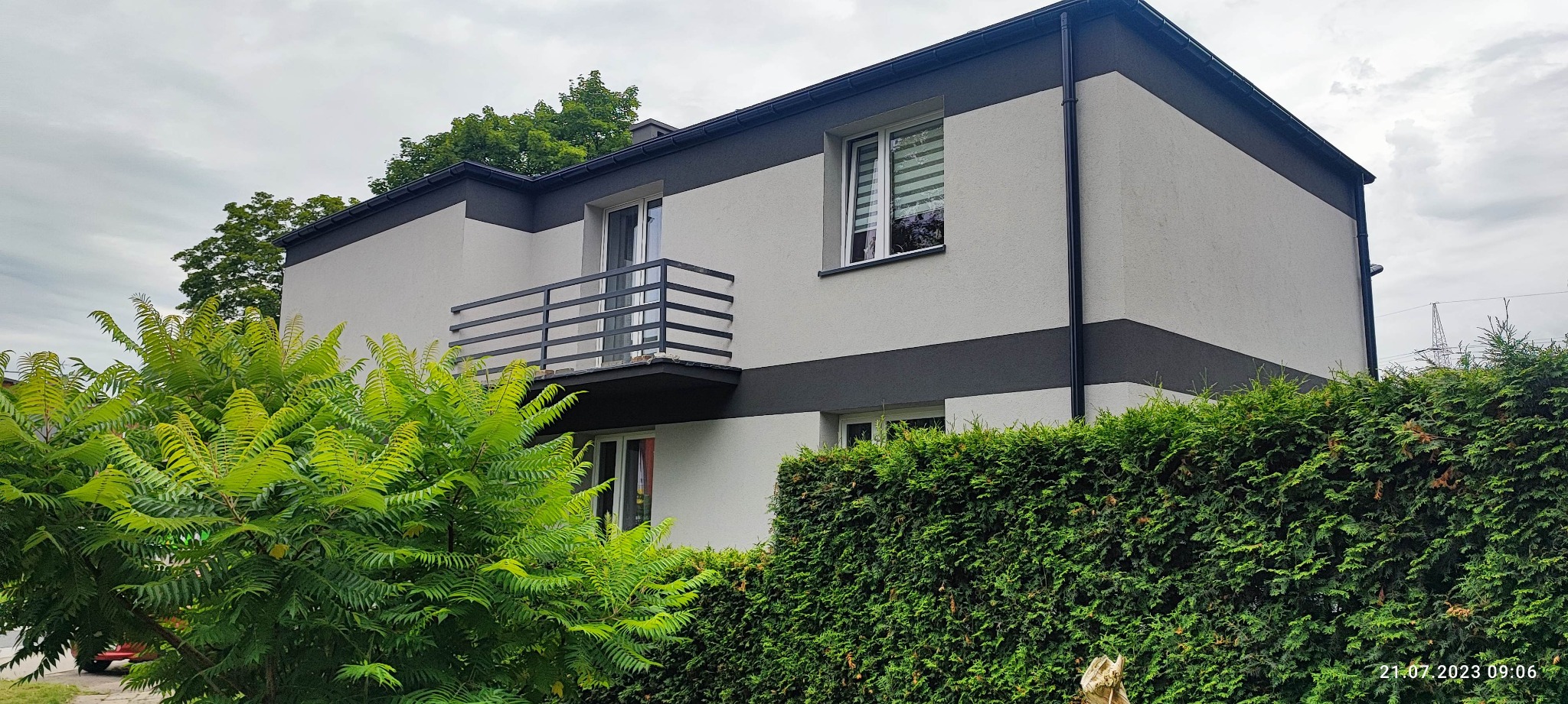 Dwukondygnacyjny dom z balkonem i elewacją w odcieniach szarości, otoczony zielenią drzew i żywopłotu.