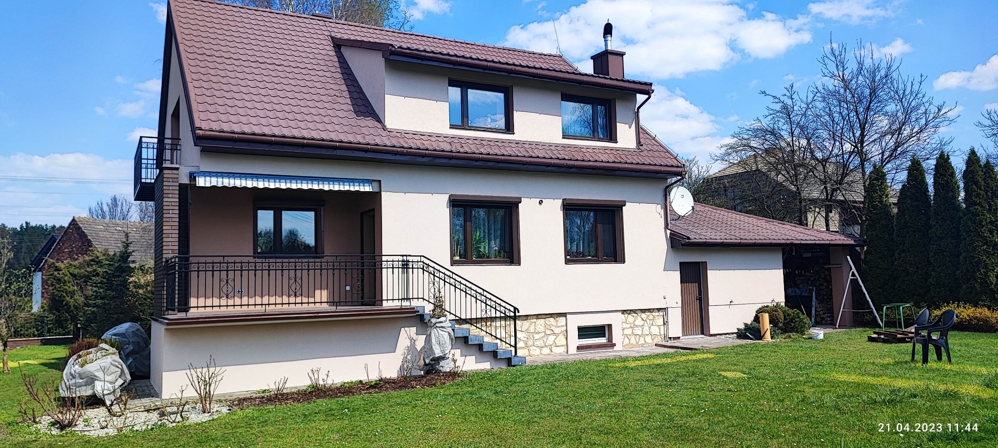 Dwukondygnacyjny dom z brązowym dachem, balkonem z kutą balustradą, oknami z roletami zewnętrznymi i zadbanym trawnikiem.