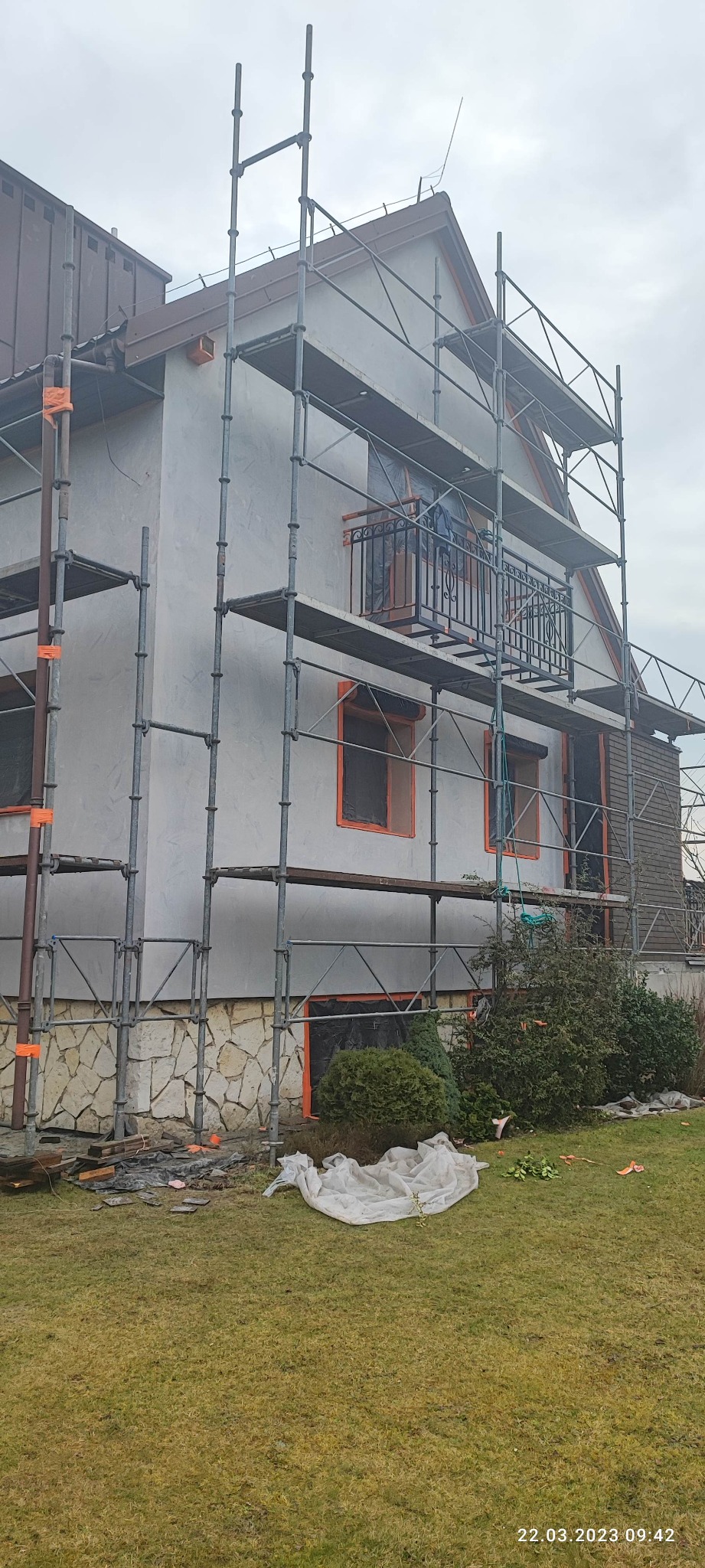 Dom jednorodzinny w trakcie ocieplania, z widocznym rusztowaniem, zabezpieczonymi oknami i fragmentem elewacji pokrytej tynkiem.