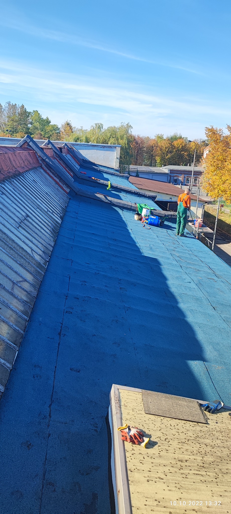 Pracownik w zielonym kombinezonie i pomarańczowej koszulce na niebieskim dachu z papy, widoczne narzędzia i puszki z farbą, w tle budynki i drzewa.