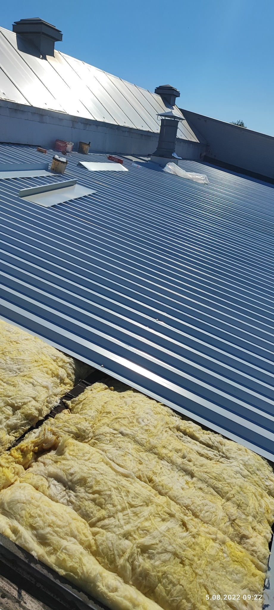 Blacha trapezowa na dachu w trakcie montażu, widoczna warstwa izolacji z wełny mineralnej, kominy wentylacyjne i akcesoria dekarskie na górze.