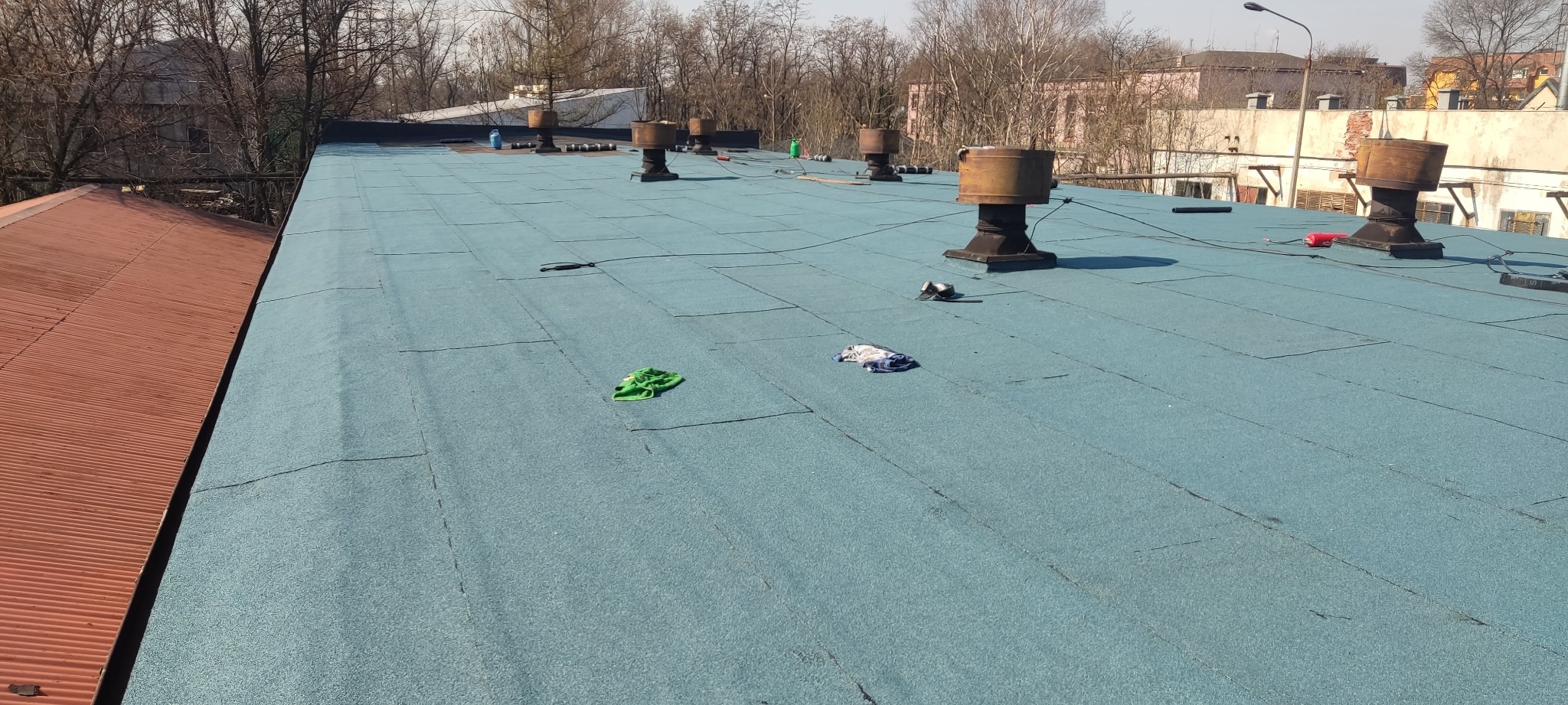 Niebieski dach pokryty papą z widocznymi kominami wentylacyjnymi, narzędziami i szmatkami w tle budynki i drzewa.