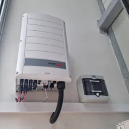 Zamontowany inwerter SolarEdge z podłączonymi przewodami oraz skrzynka rozdzielcza DC Noark na białej ścianie z widoczną konstrukcją stalową i naklejką ostrzegawczą.