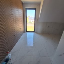 Łukasz Hyrchel - Wykończone wnętrze poddasza z drewnianą boazerią i białymi płytkami imitującymi marmur na podłodze i ścianach. Widok na zewnątrz przez okno.