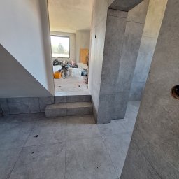 Łukasz Hyrchel - Szare, matowe płytki podłogowe i ścienne, układane w nowoczesnym wnętrzu w trakcie prac wykończeniowych. Widoczne schody i fragment okna w tle.