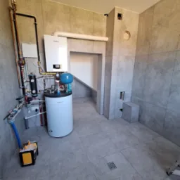 Szara łazienka z zamontowanym piecem gazowym Vaillant, instalacją hydrauliczną i wentylacją. Widoczny odpływ podłogowy i skrzynka narzędziowa.