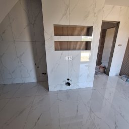 Łukasz Hyrchel - Wykończona łazienka z białymi płytkami imitującymi marmur, drewniane akcenty w niszach. Widoczne przyłącza hydrauliczne i elektryczne. Otwór drzwiowy w tle.