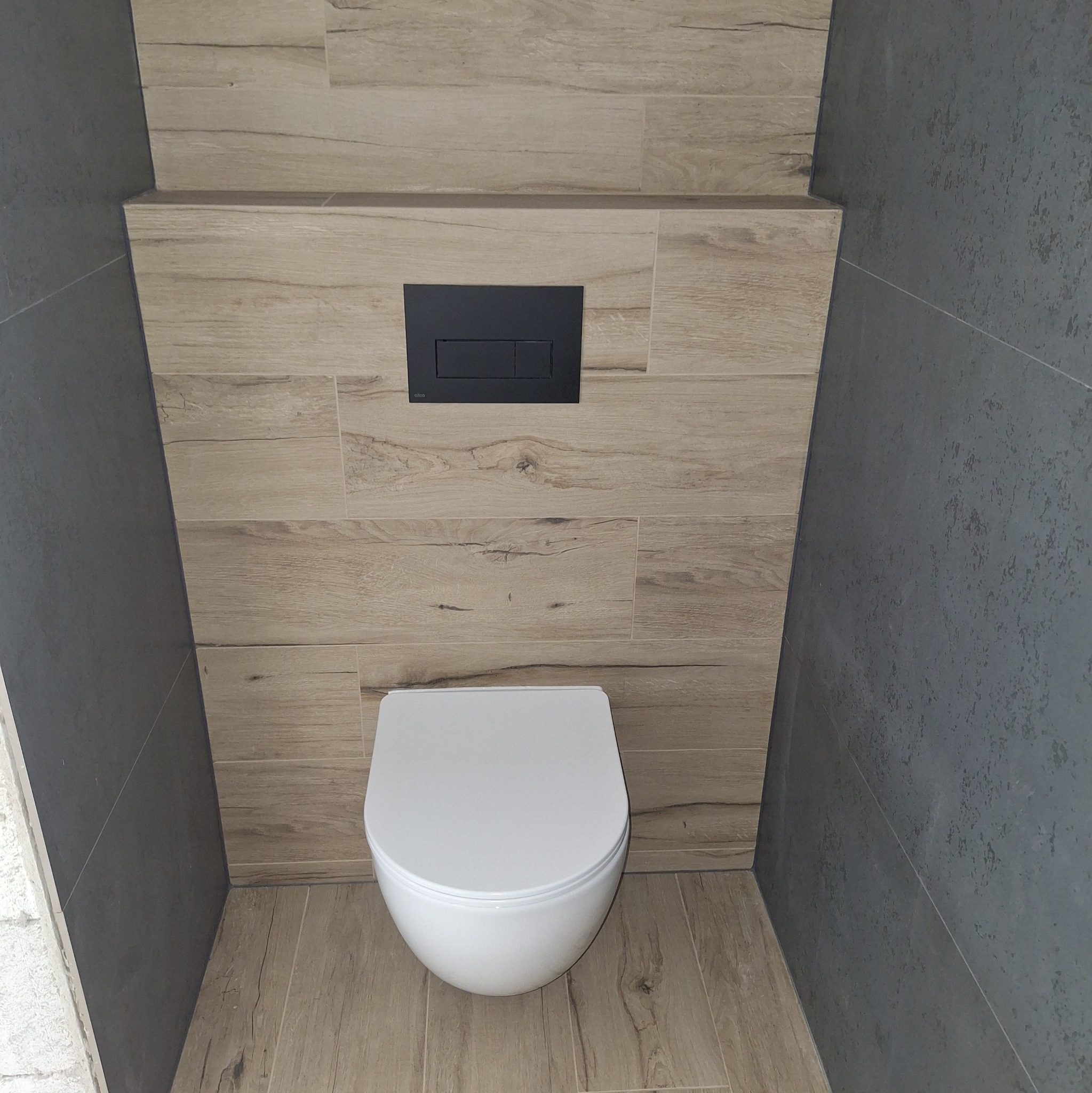 Nowoczesna toaleta podwieszana z czarnym przyciskiem spłukującym, obudowana płytkami imitującymi drewno i szarym gresem. Minimalistyczny design.