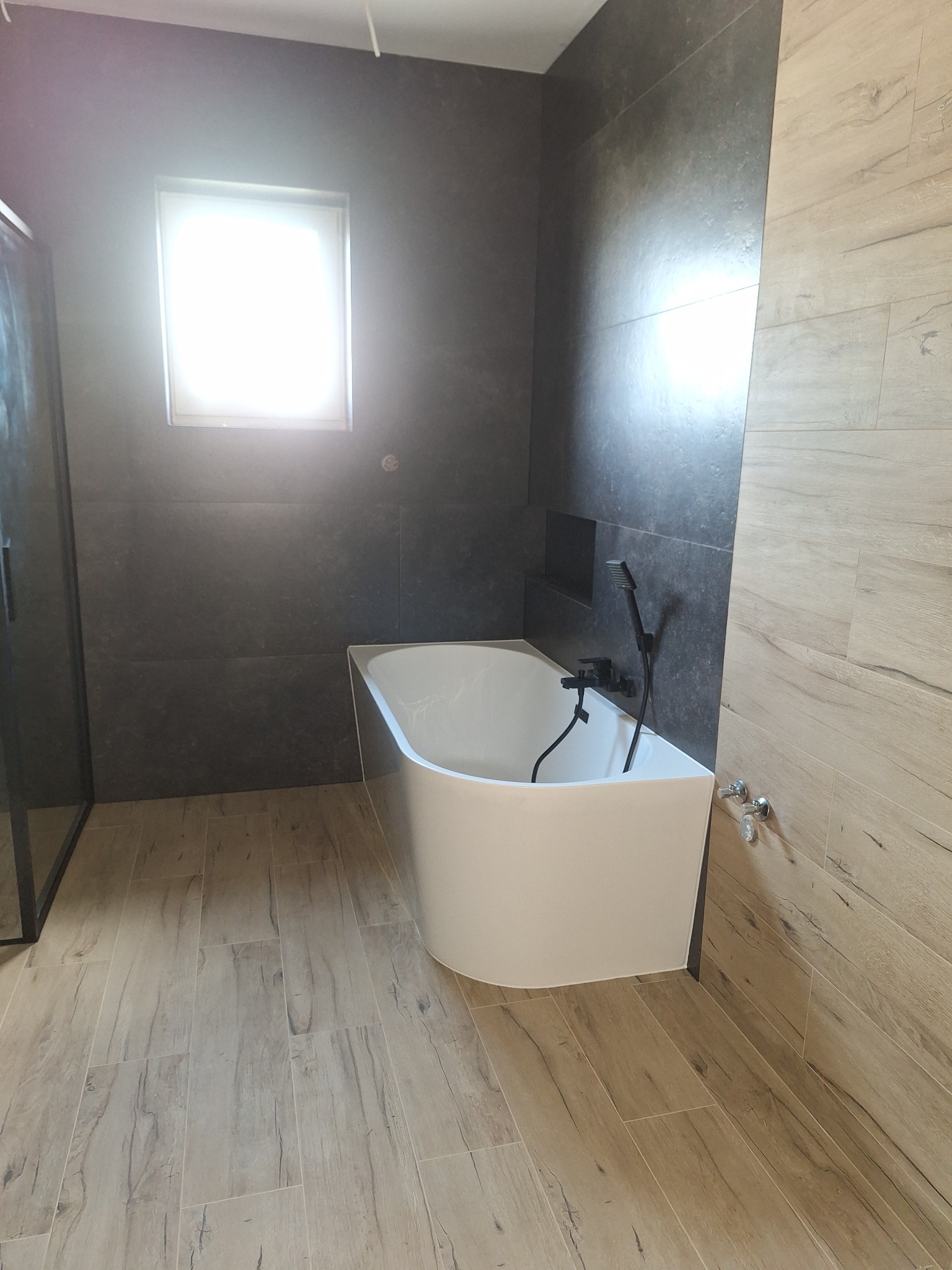 Nowoczesna łazienka z wanną wolnostojącą, czarną armaturą i płytkami imitującymi drewno na podłodze i ścianie. Minimalistyczny design, okno w tle.