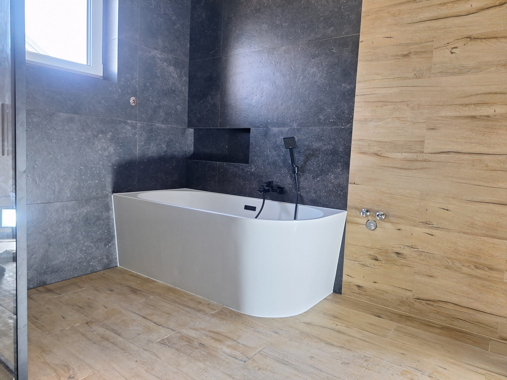 Nowoczesna łazienka: biała wanna z czarną armaturą na tle grafitowych płytek i drewnopodobnej okładziny, jasna podłoga, minimalistyczny design.