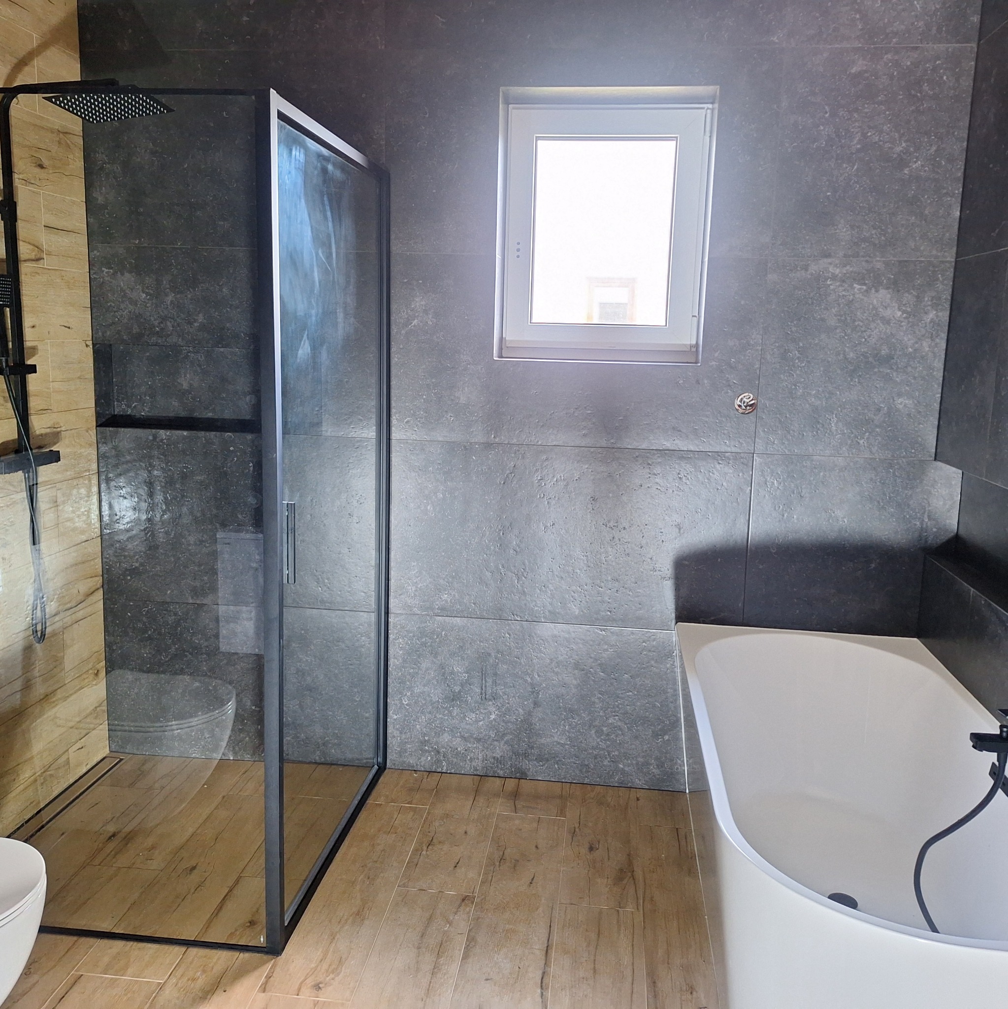 Nowoczesna łazienka z kabiną typu walk-in, drewnopodobnymi płytkami podłogowymi i szarymi płytkami ściennymi. Widoczne okno i wanna.