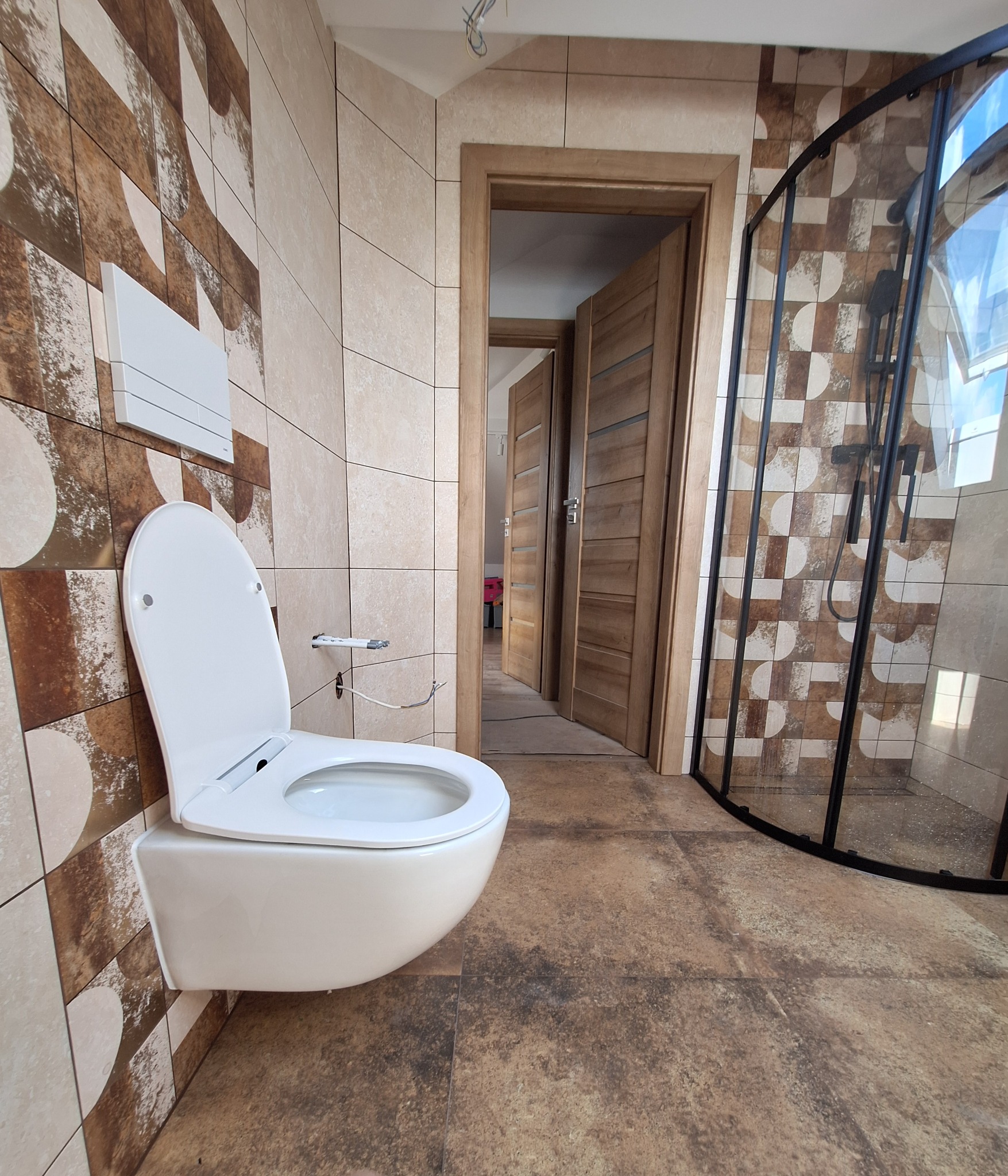 Nowoczesna łazienka po remoncie: podwieszana toaleta, kabina prysznicowa z czarnymi profilami i płytki w odcieniach beżu i brązu z geometrycznym wzorem.