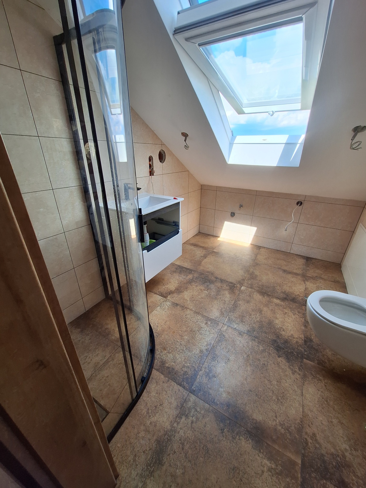 Wykończona łazienka na poddaszu z beżowymi kaflami i brązową podłogą. Widoczna kabina prysznicowa z czarnymi ramami, umywalka i toaleta. Otwarty świetlik wpuszcza światło.