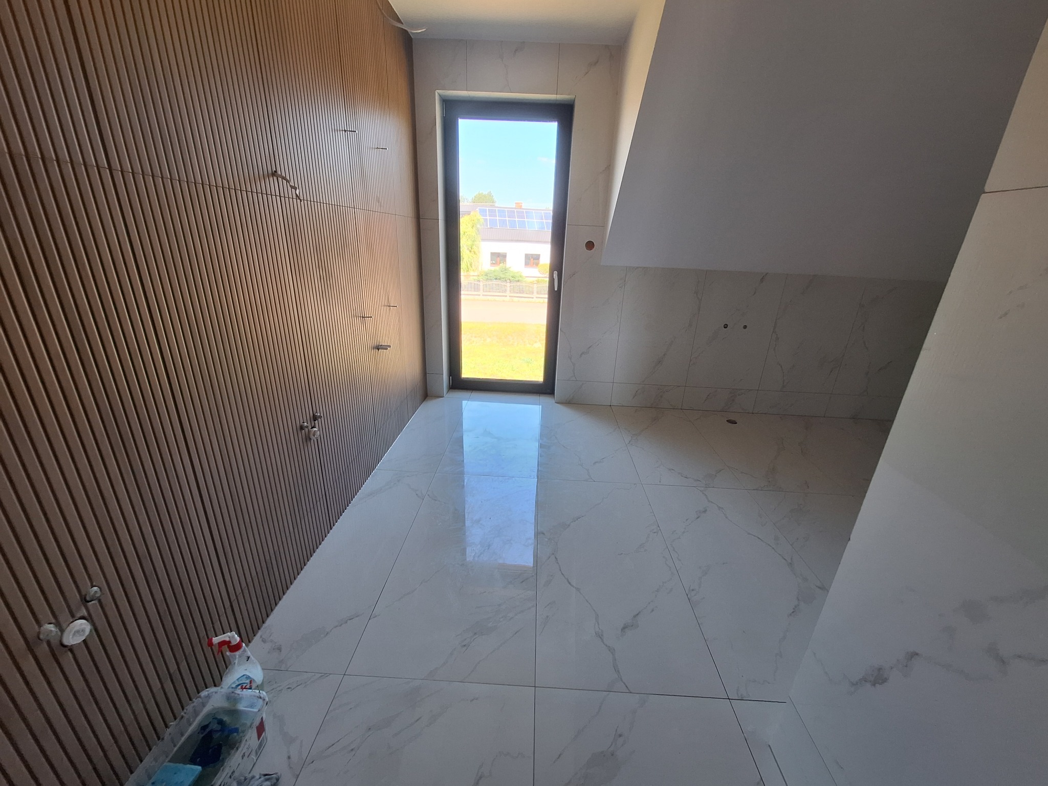 Wykończone wnętrze poddasza z drewnianą boazerią i białymi płytkami imitującymi marmur na podłodze i ścianach. Widok na zewnątrz przez okno.