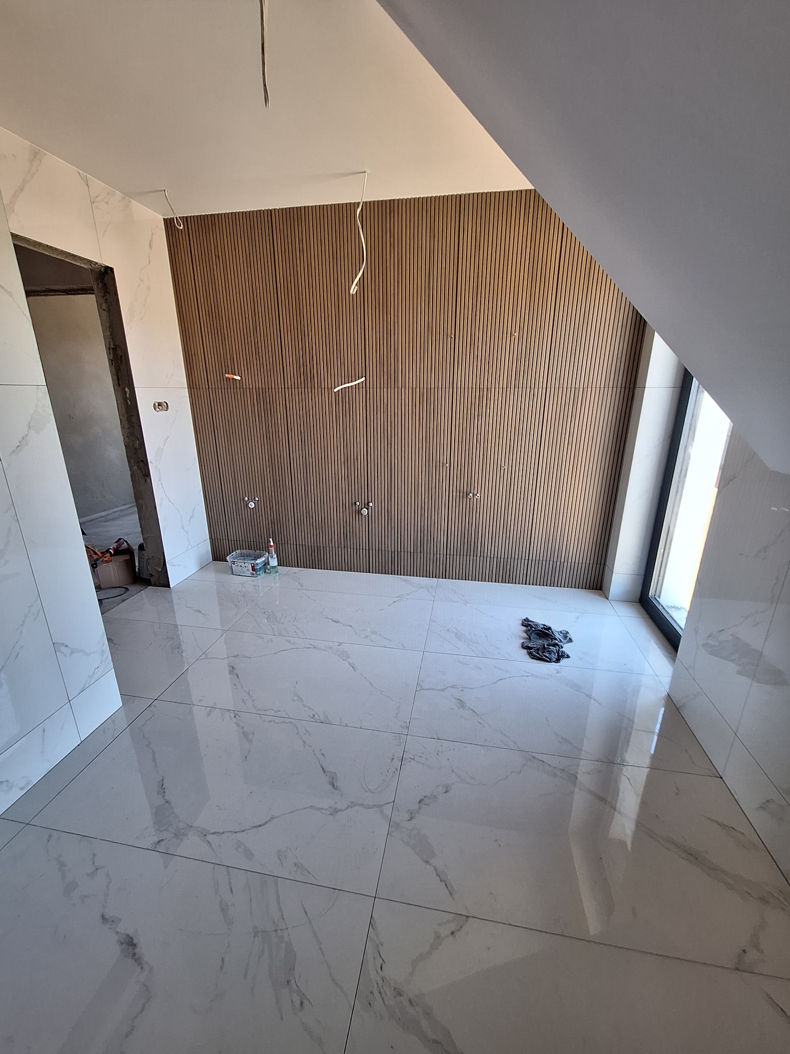 Wykończona łazienka na poddaszu w Mielcu: białe płytki imitujące marmur na podłodze i ścianach, drewniane panele ścienne, okno dachowe. Widoczne przygotowania do montażu armatury.