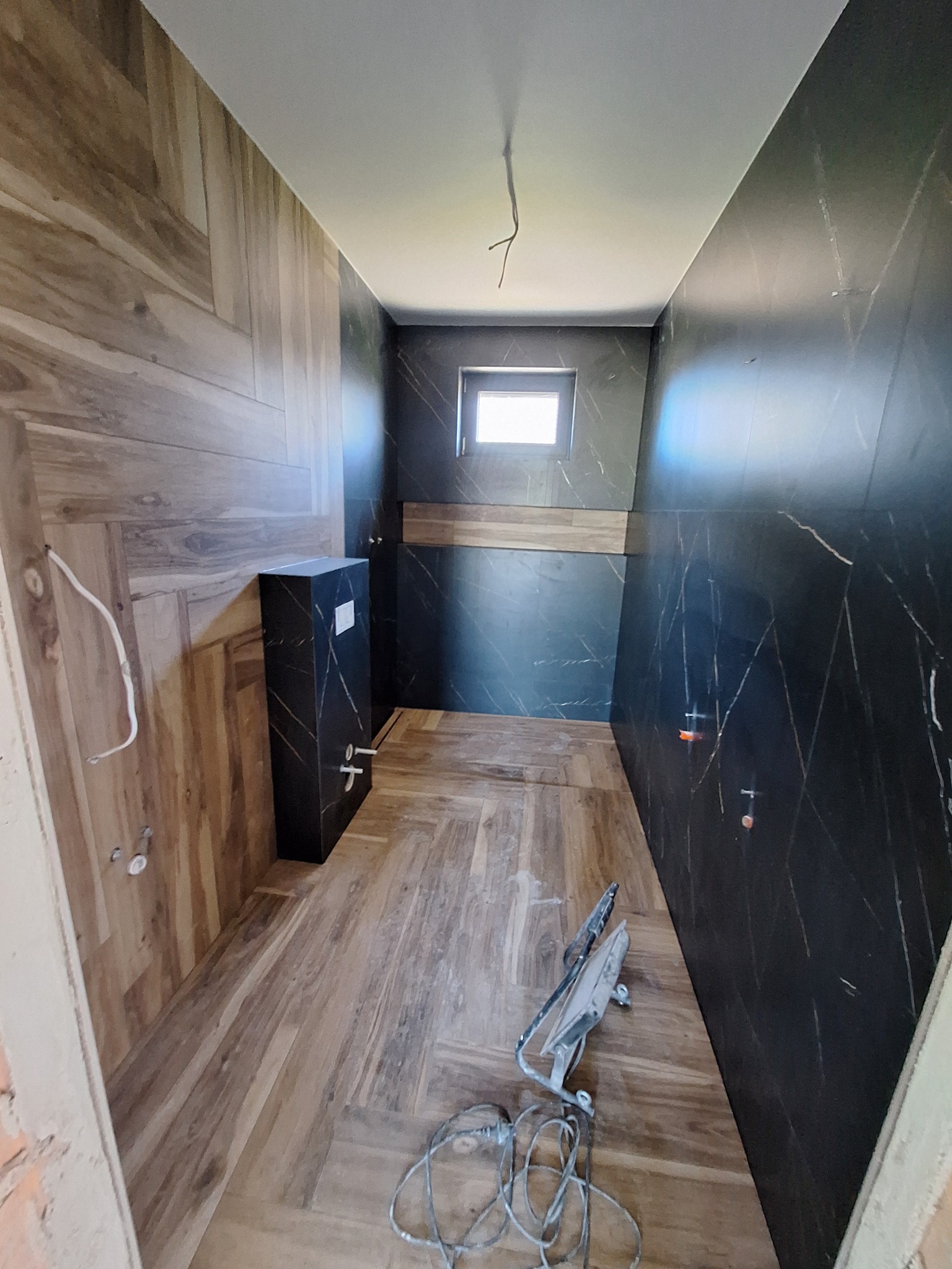 Wnętrze łazienki w trakcie prac wykończeniowych: ułożone płytki imitujące drewno na podłodze i ścianie, czarne płytki z marmurowym wzorem na pozostałych ścianach.