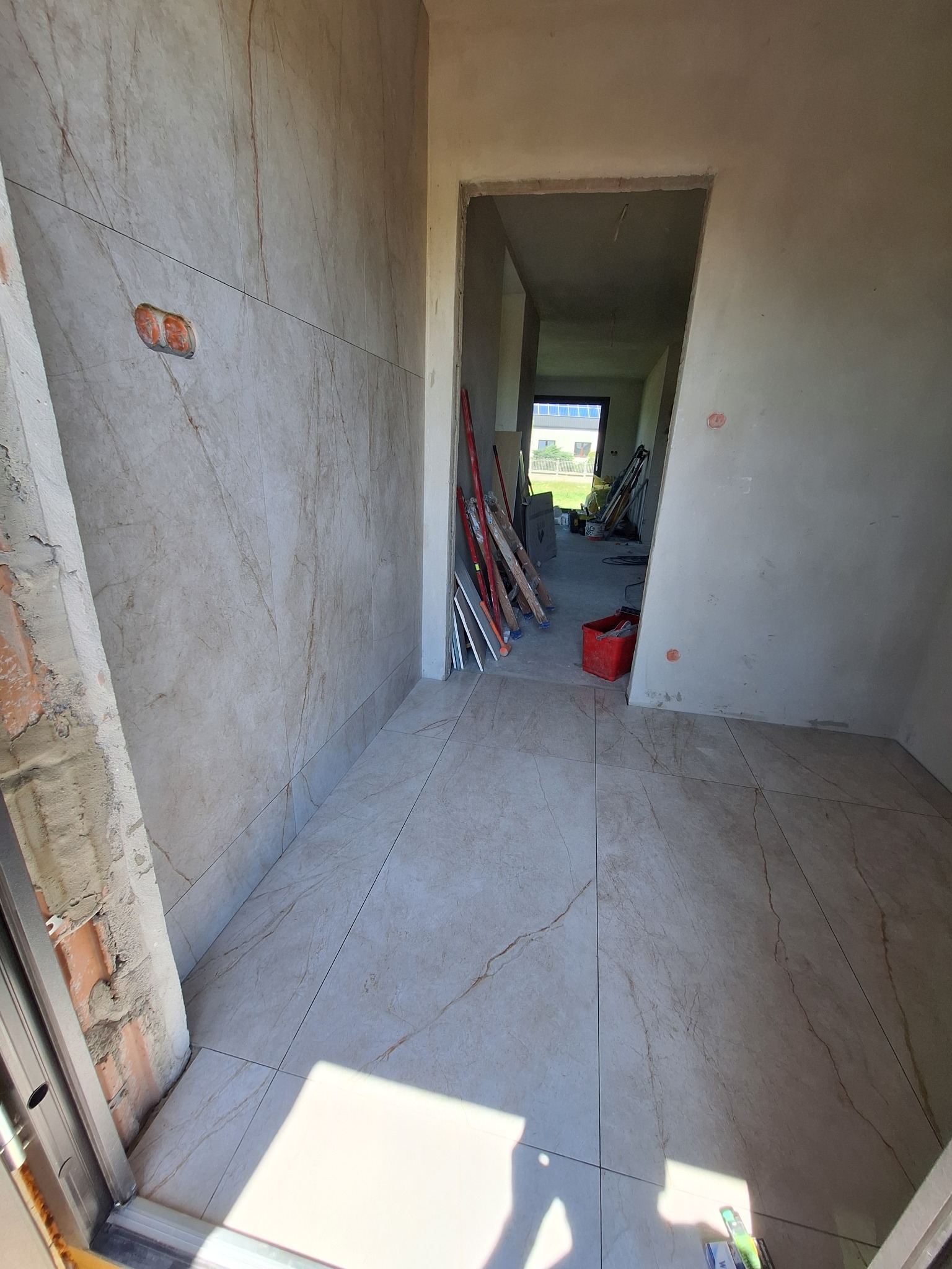 Wnętrze w trakcie prac: ułożone płytki podłogowe i ścienne imitujące marmur. Widoczne narzędzia, surowe ściany i otwór drzwiowy w stanie wykończeniowym.