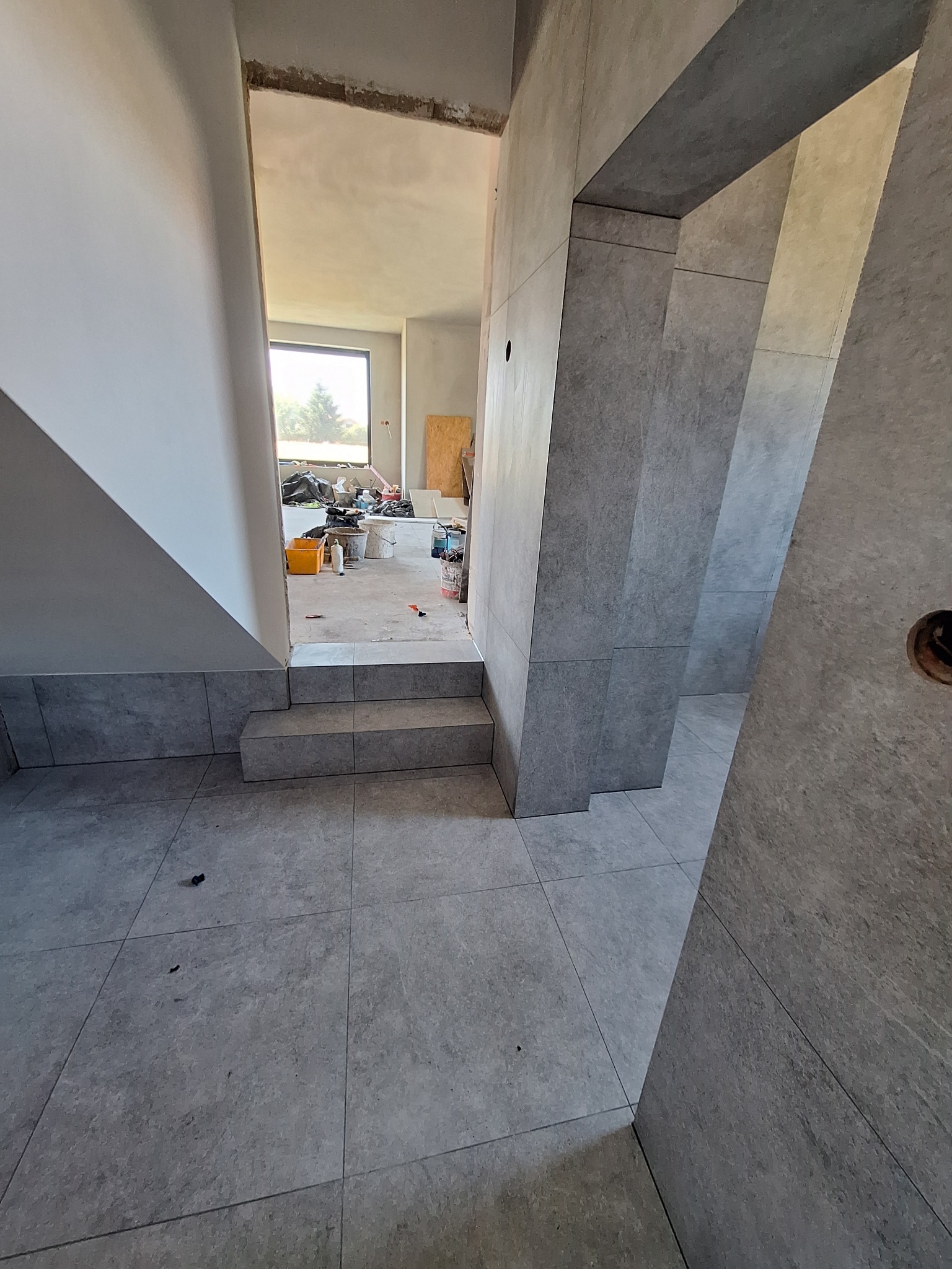 Szare, matowe płytki podłogowe i ścienne, układane w nowoczesnym wnętrzu w trakcie prac wykończeniowych. Widoczne schody i fragment okna w tle.